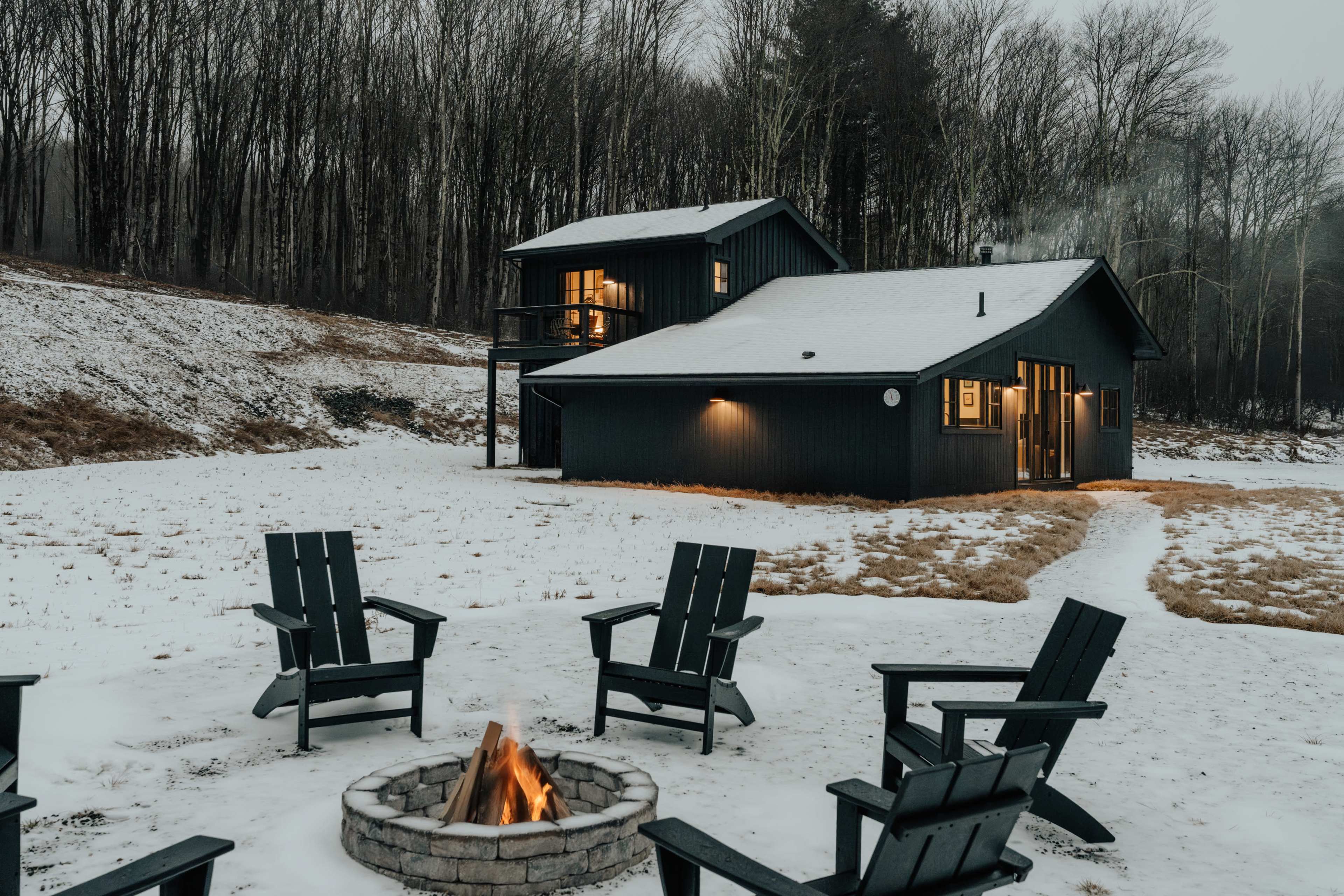 A black two-story cabin with large windows is surrounded by a snowy landscape and a fire pit with four Adirondack chairs arranged around it.