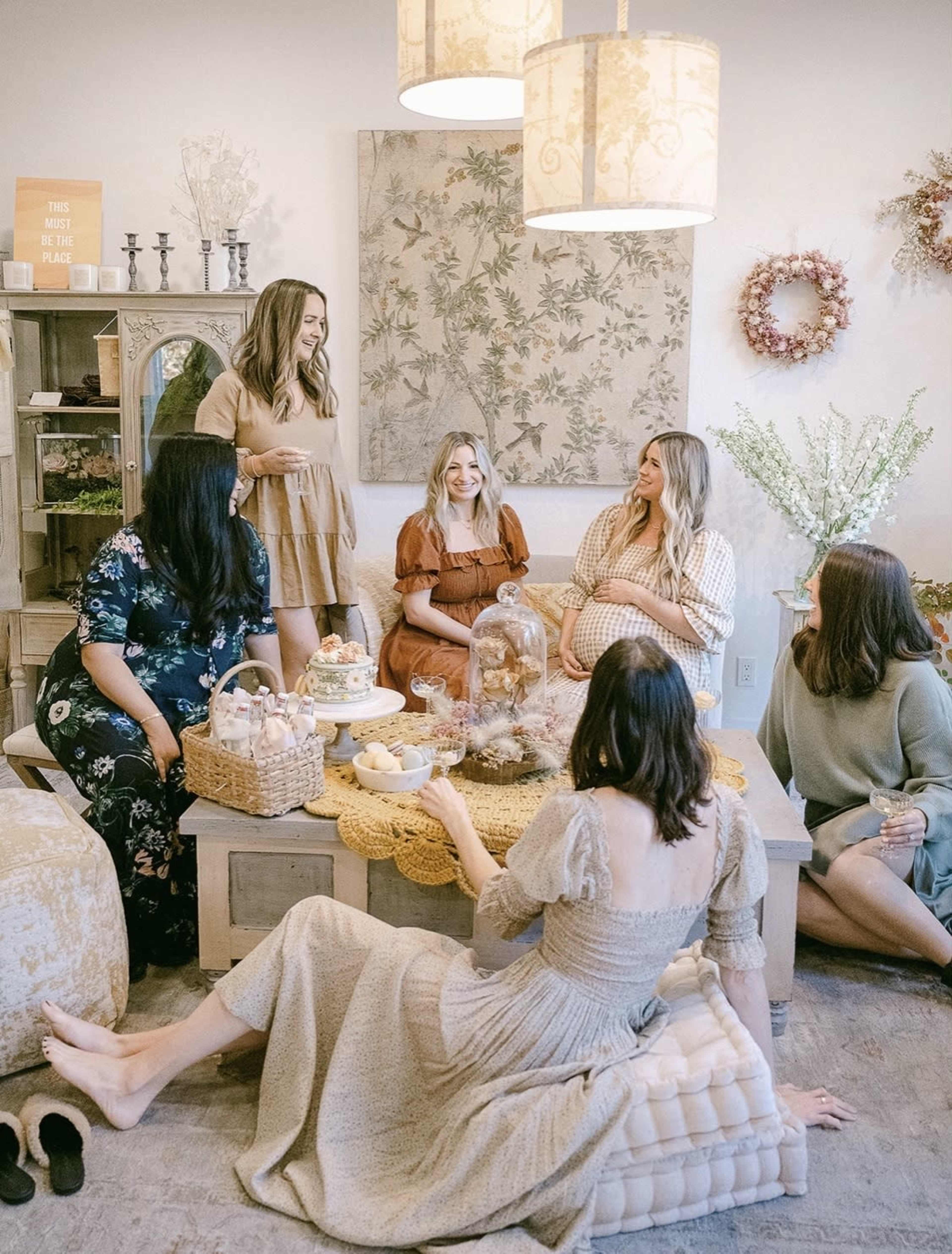 A group of seven women sits together in a cozy indoor setting, enjoying a gathering with a table filled with food and drinks.