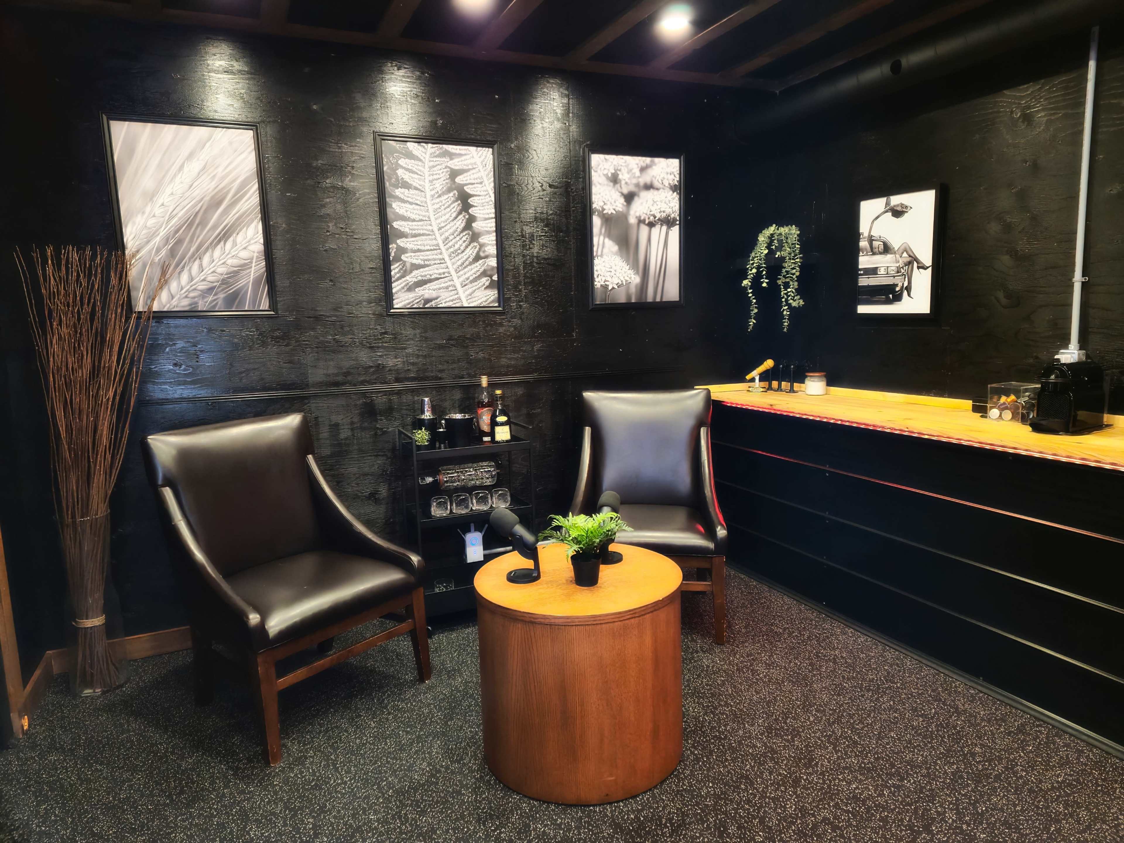 The image depicts a cozy corner with two black armchairs and a small round table, surrounded by black walls adorned with framed black-and-white photographs.