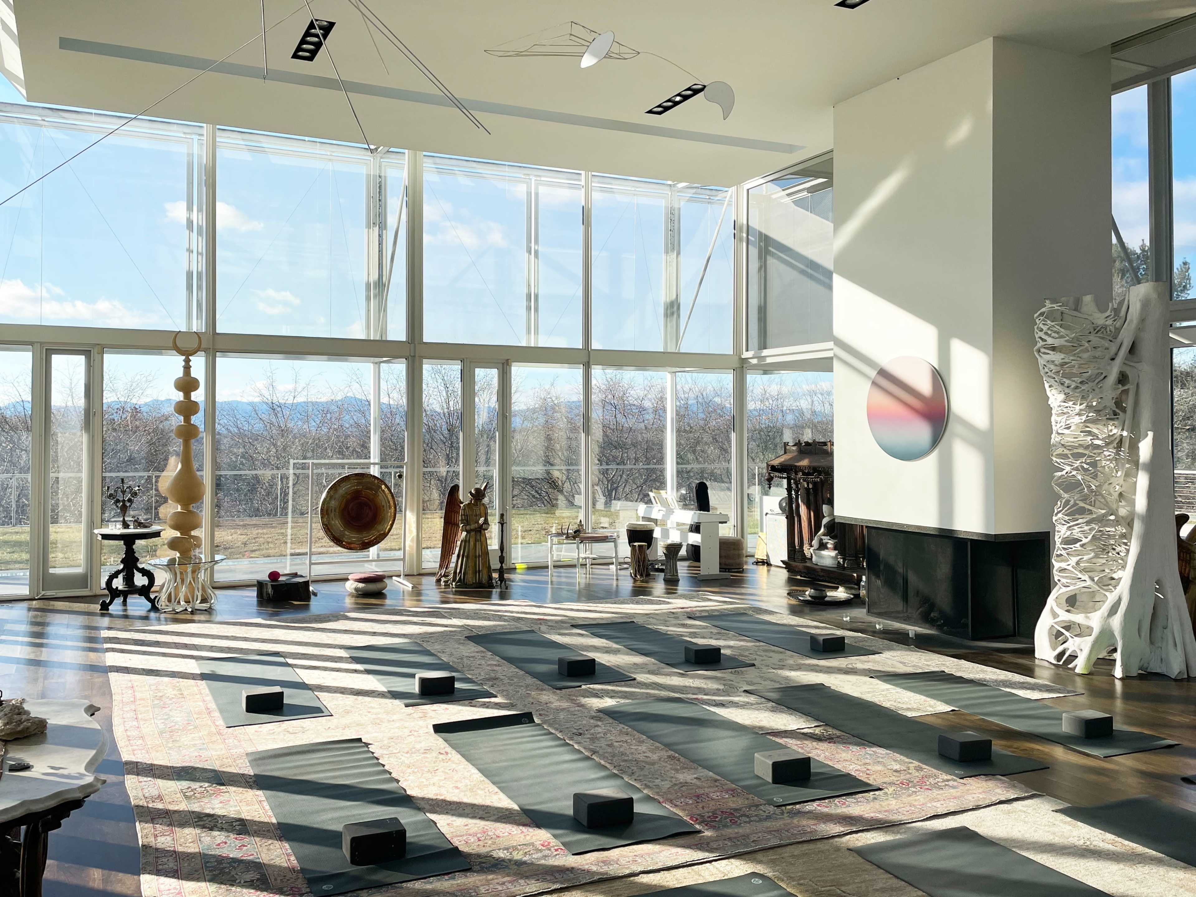 A spacious yoga studio with large windows, wooden flooring, and yoga mats neatly arranged on the ground, along with various art pieces and decorative objects.