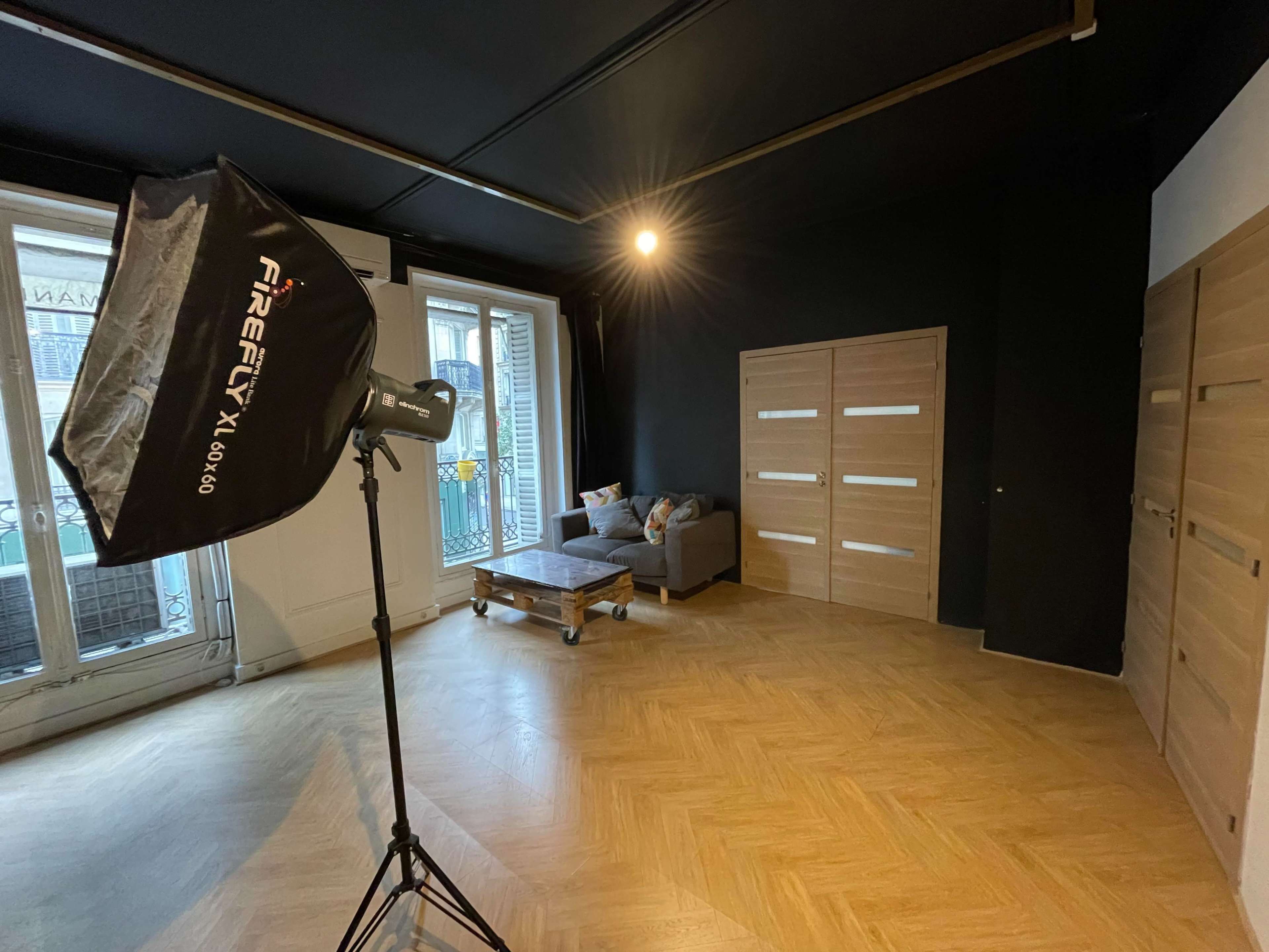 A spacious room featuring a sofa on a wooden pallet, a photography light setup, and two wooden doors against a black wall.