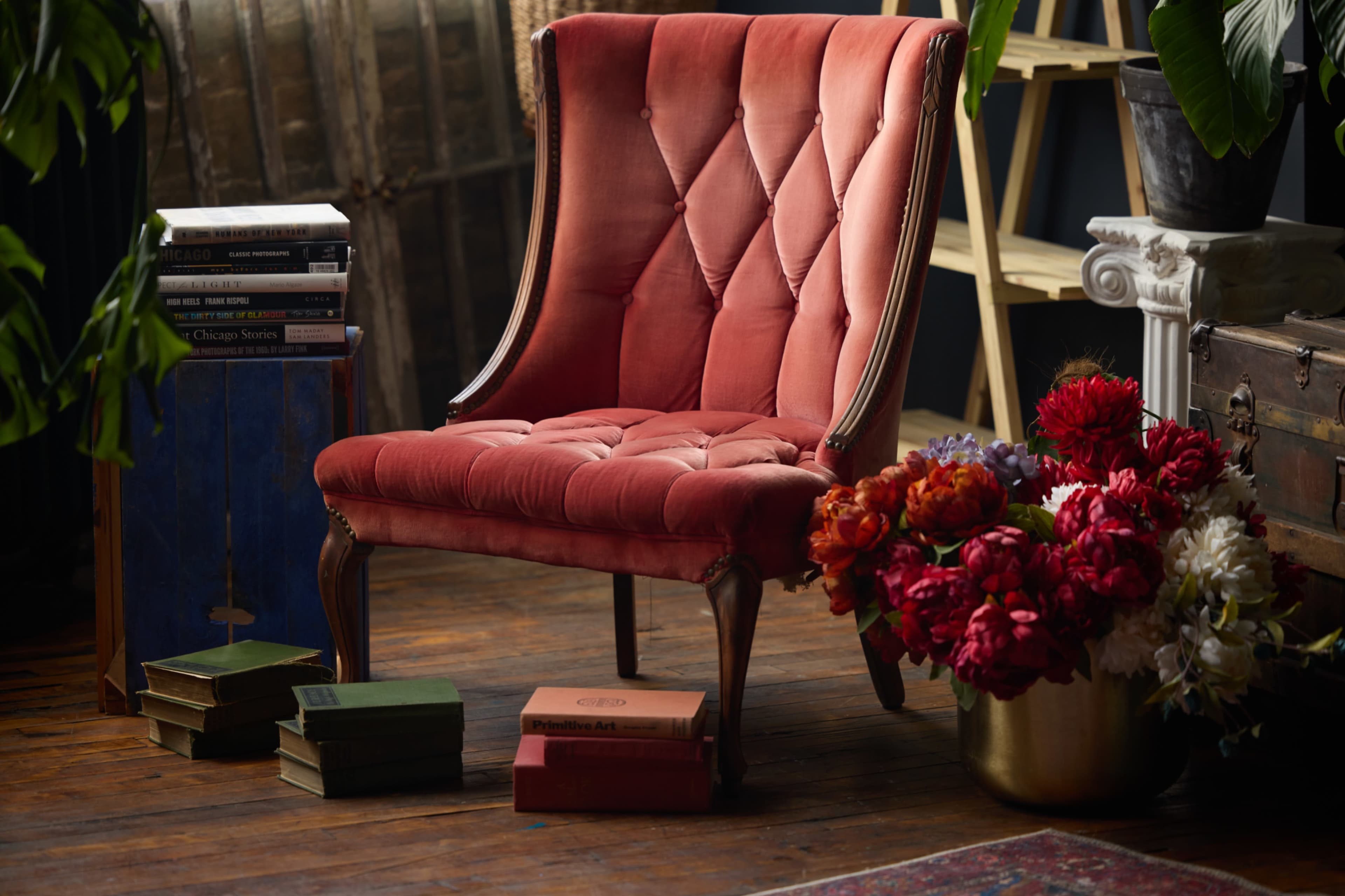A vintage red upholstered chair is positioned next to a potted plant and a bouquet of various flowers, with stacks of books nearby on a wooden floor.