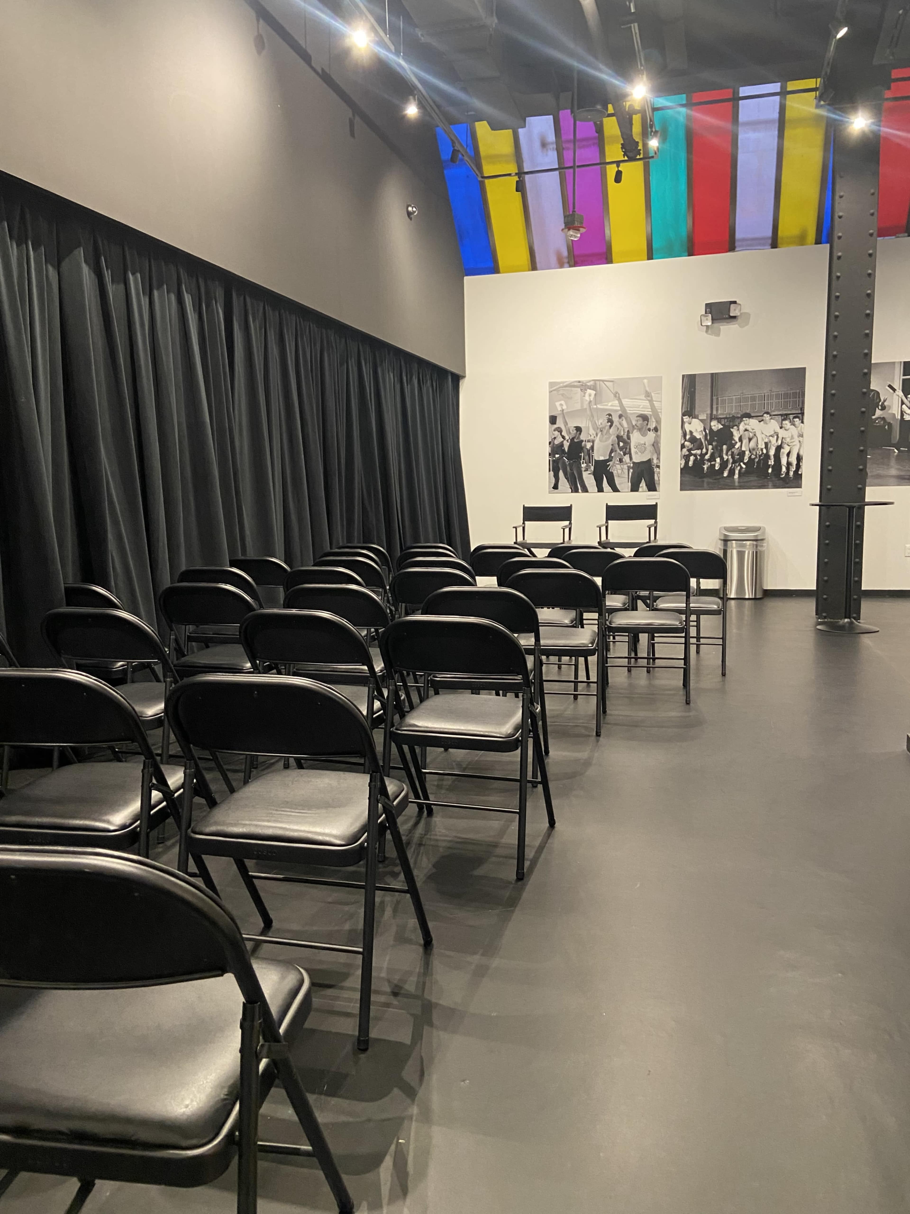A row of black folding chairs is set up facing a wall with black curtains and photographs in a well-lit room.