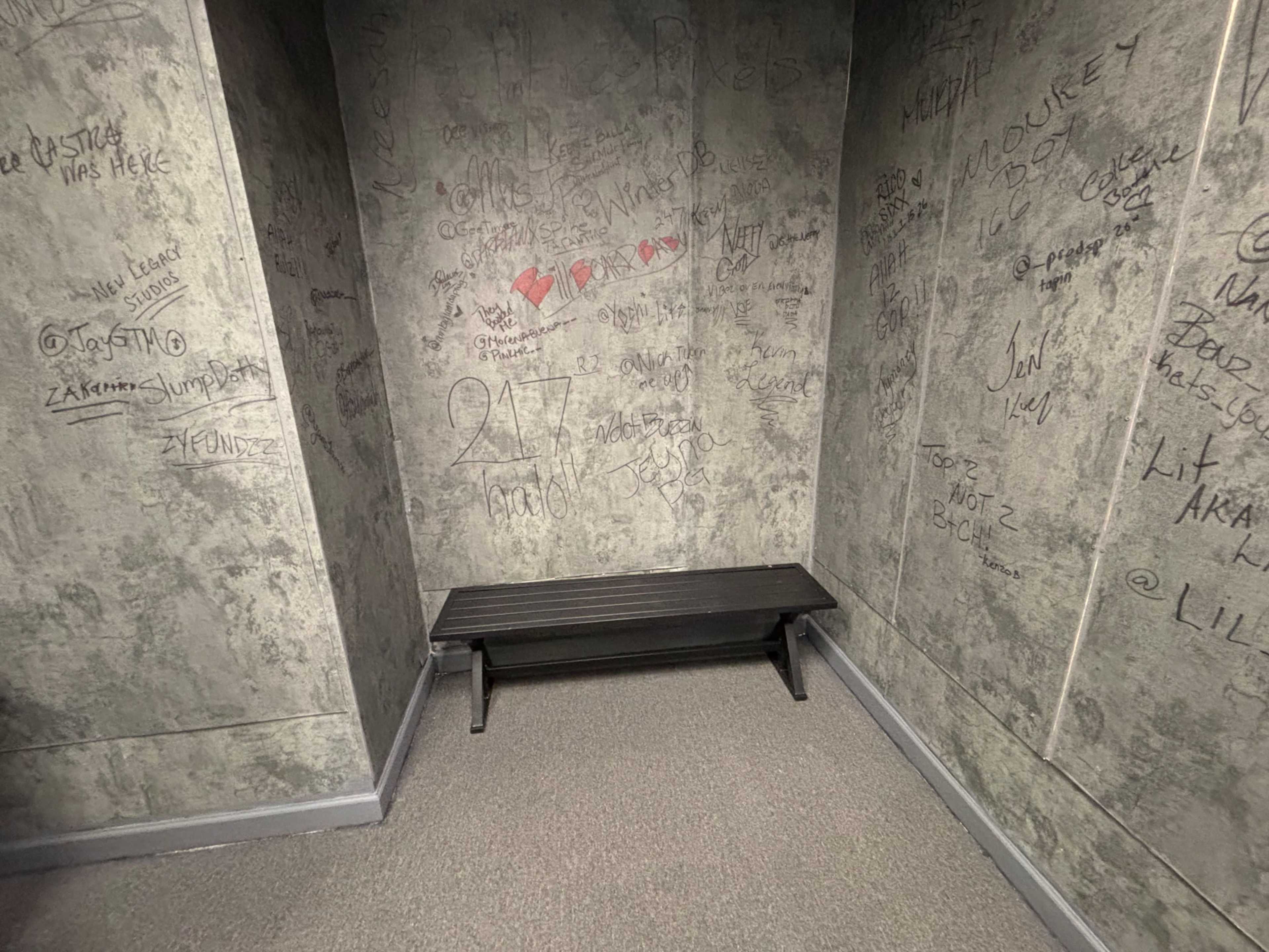 The image shows a small, empty corner of a room with a bench against a wall covered in graffiti.