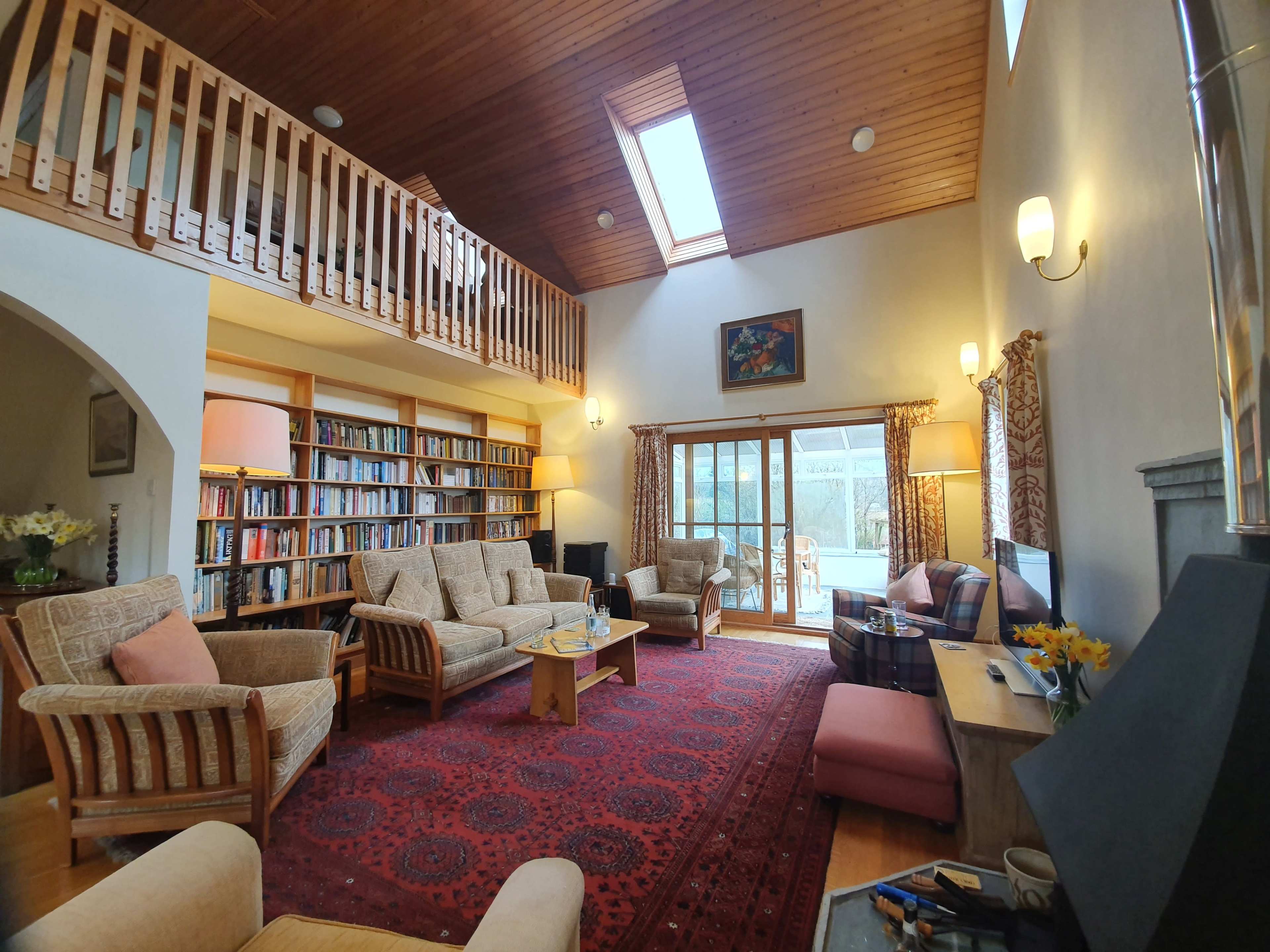 The living room features a high ceiling with wooden beams, a large bookshelf, several upholstered chairs arranged around a coffee table, and sliding glass doors leading to an outdoor area.