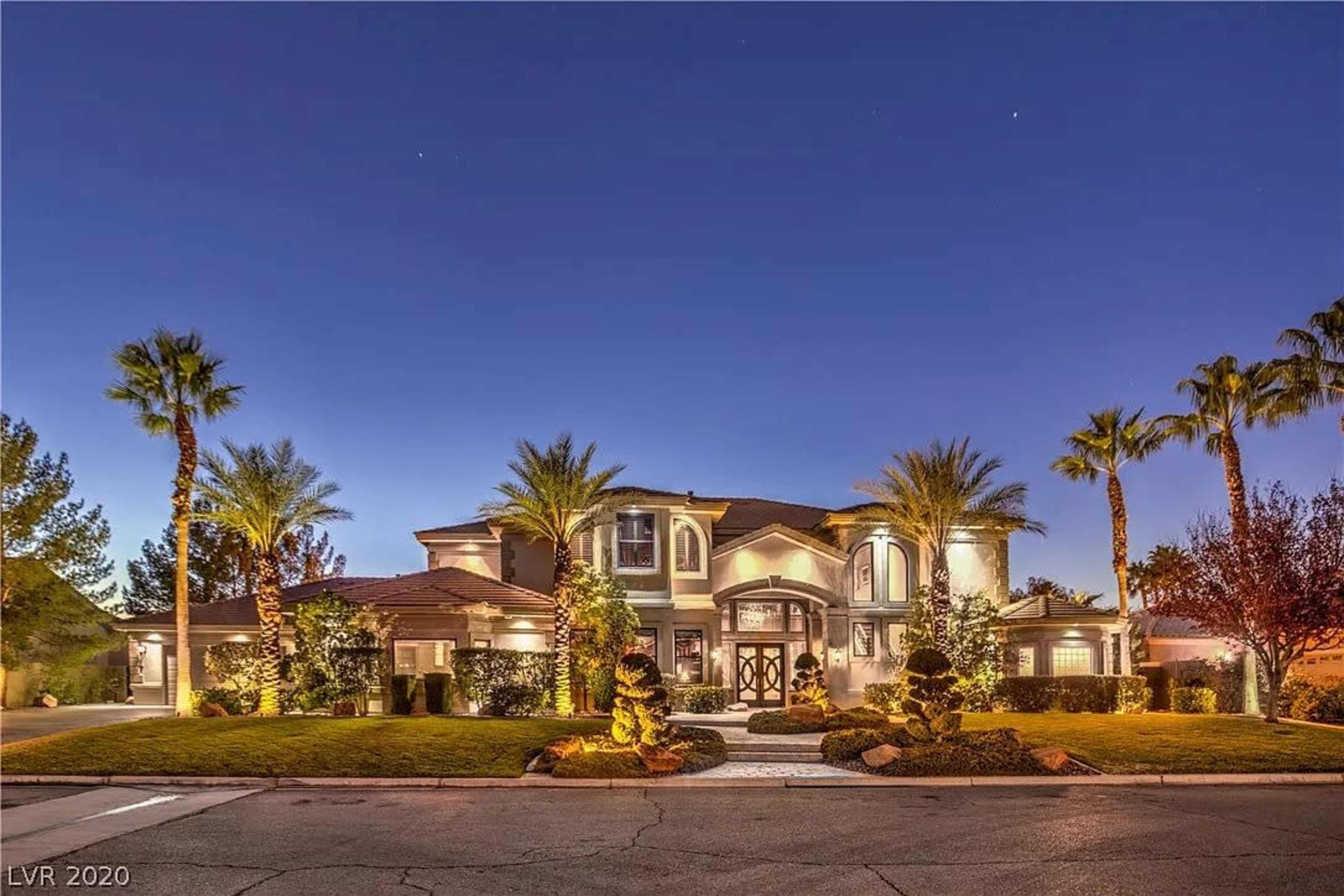 The image shows a large, illuminated two-story house surrounded by palm trees and landscaped grounds at dusk.
