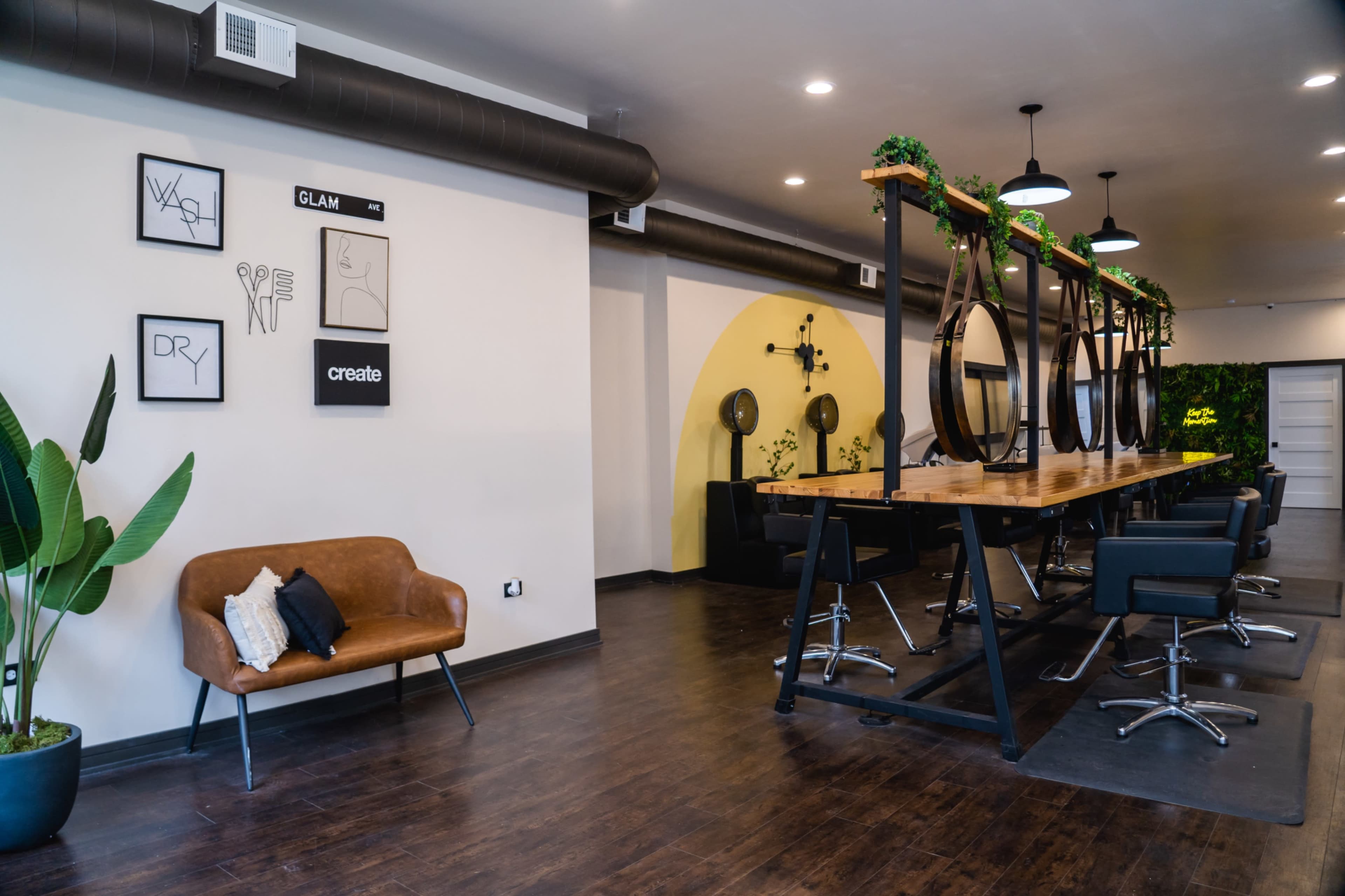 A modern salon interior with a long wooden table, black chairs, large round mirrors, and greenery on the walls.
