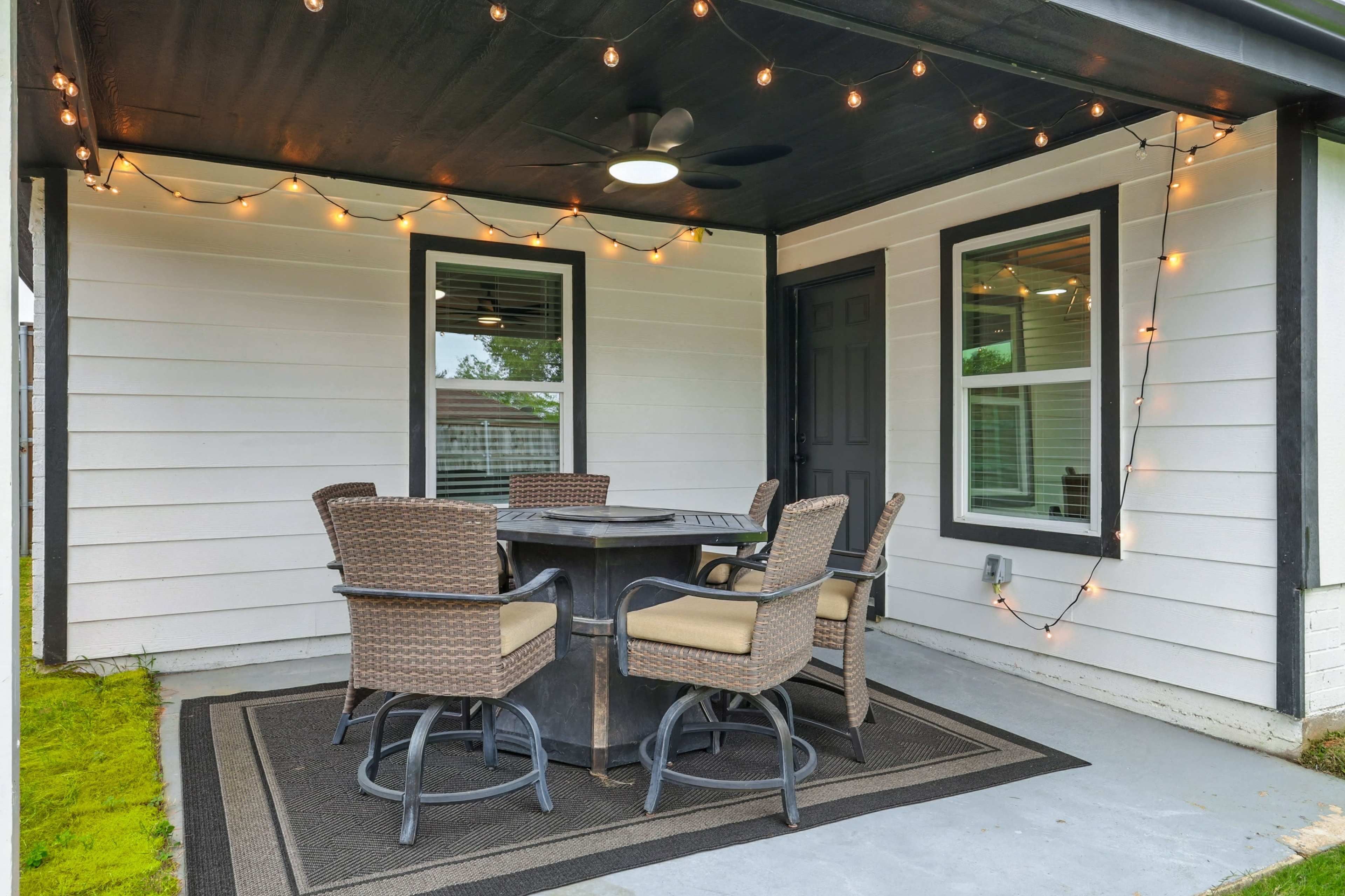 A covered porch features a round dining table surrounded by six chairs, illuminated by string lights above.