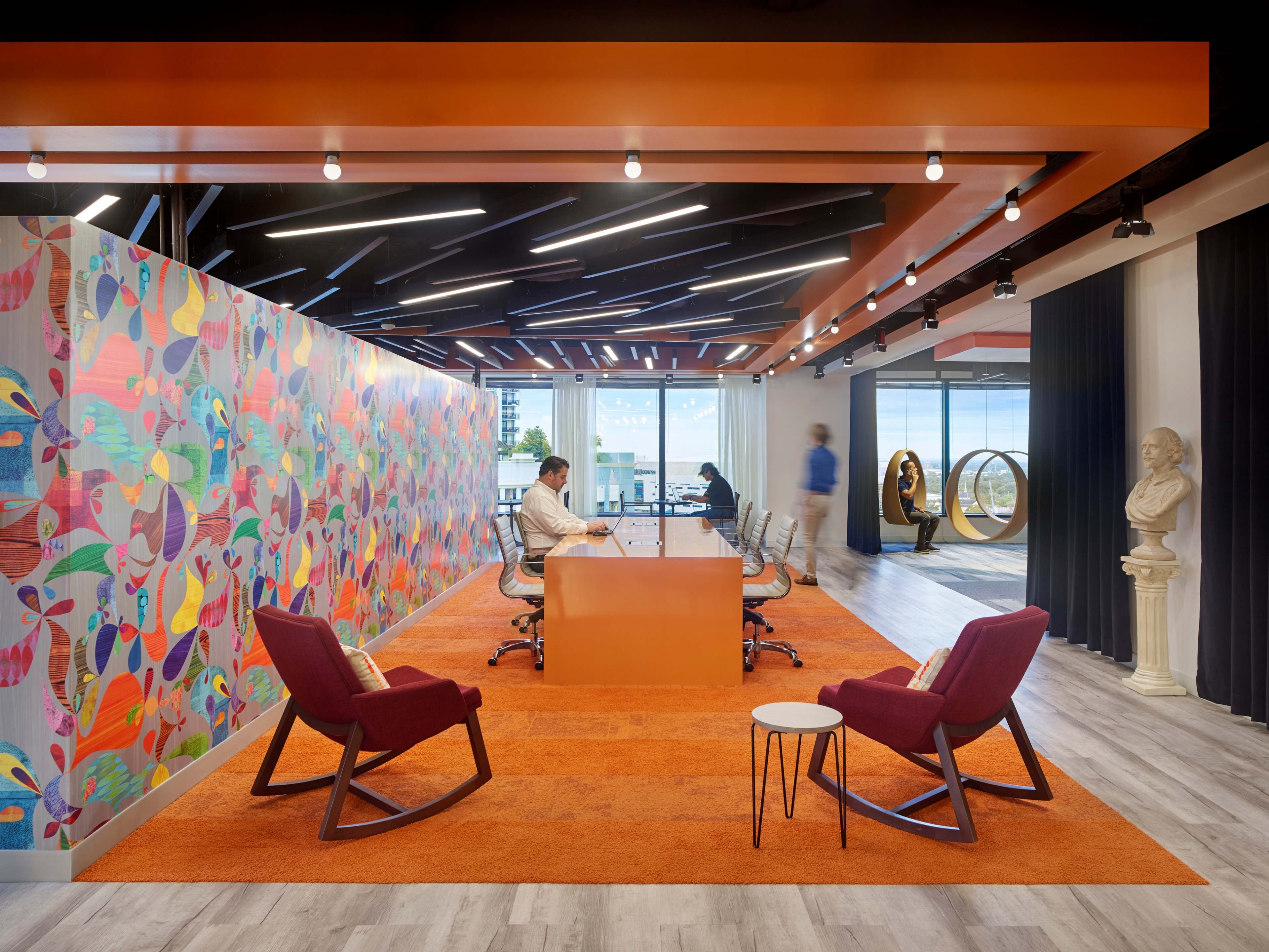 The image shows a modern office space with a colorful mural, a large table surrounded by chairs, and seating areas featuring rocking chairs and a small table.