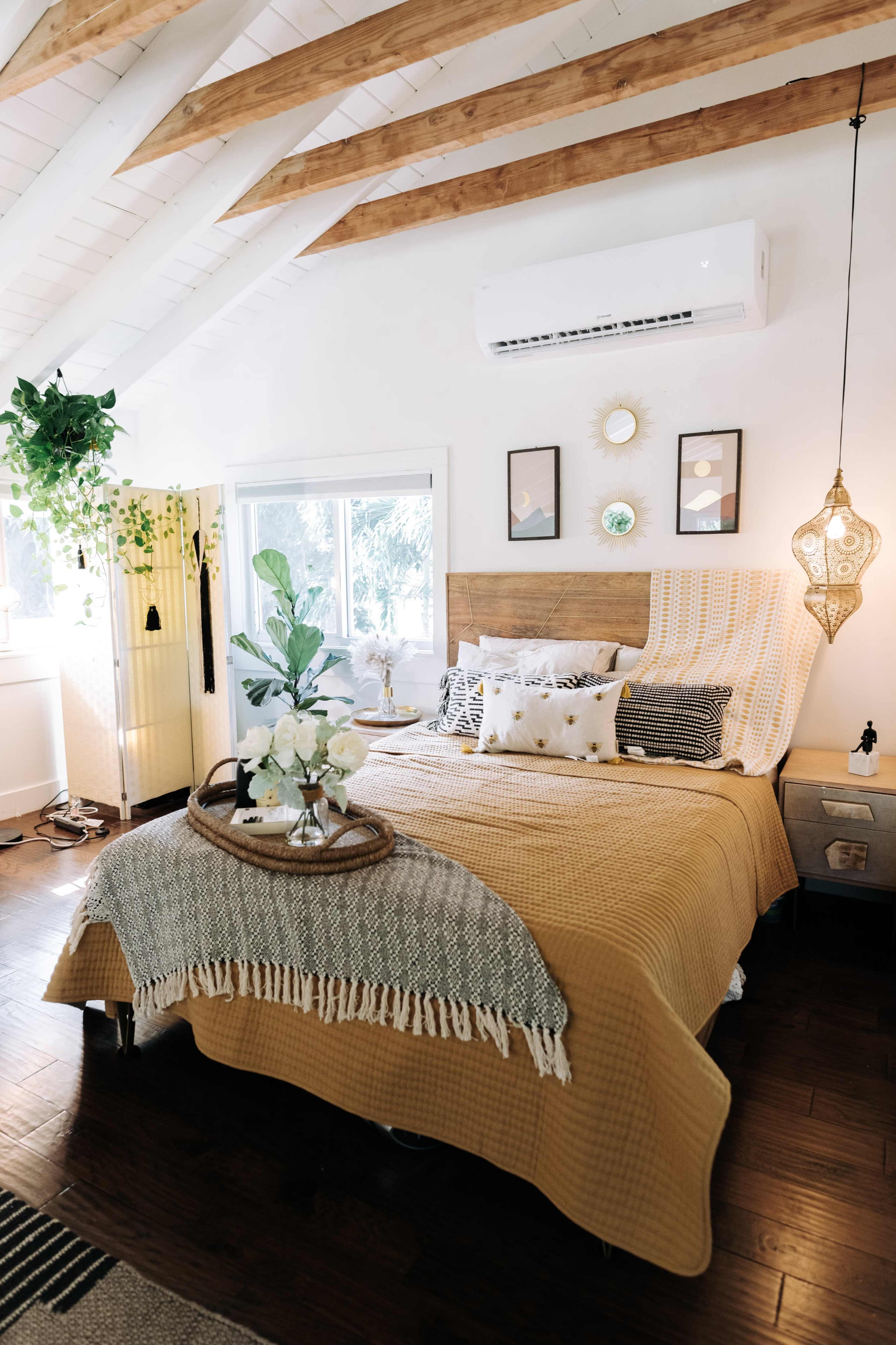 The image shows a well-decorated bedroom featuring a large bed with a yellow coverlet, surrounded by tasteful decor including plants, artwork, and a decorative light fixture.