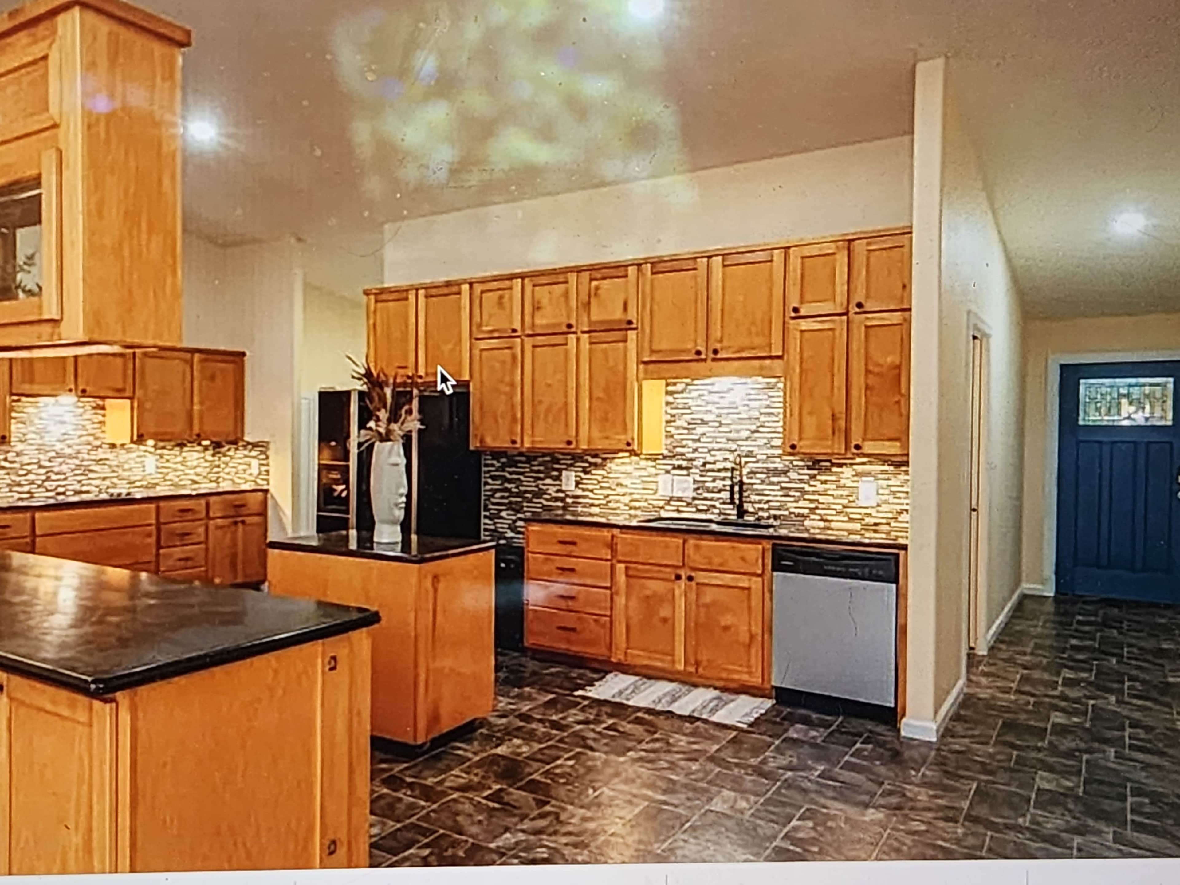 A modern kitchen with wooden cabinets, a dark countertop, and a tiled backsplash, featuring an island in the center and a blue door in the background.