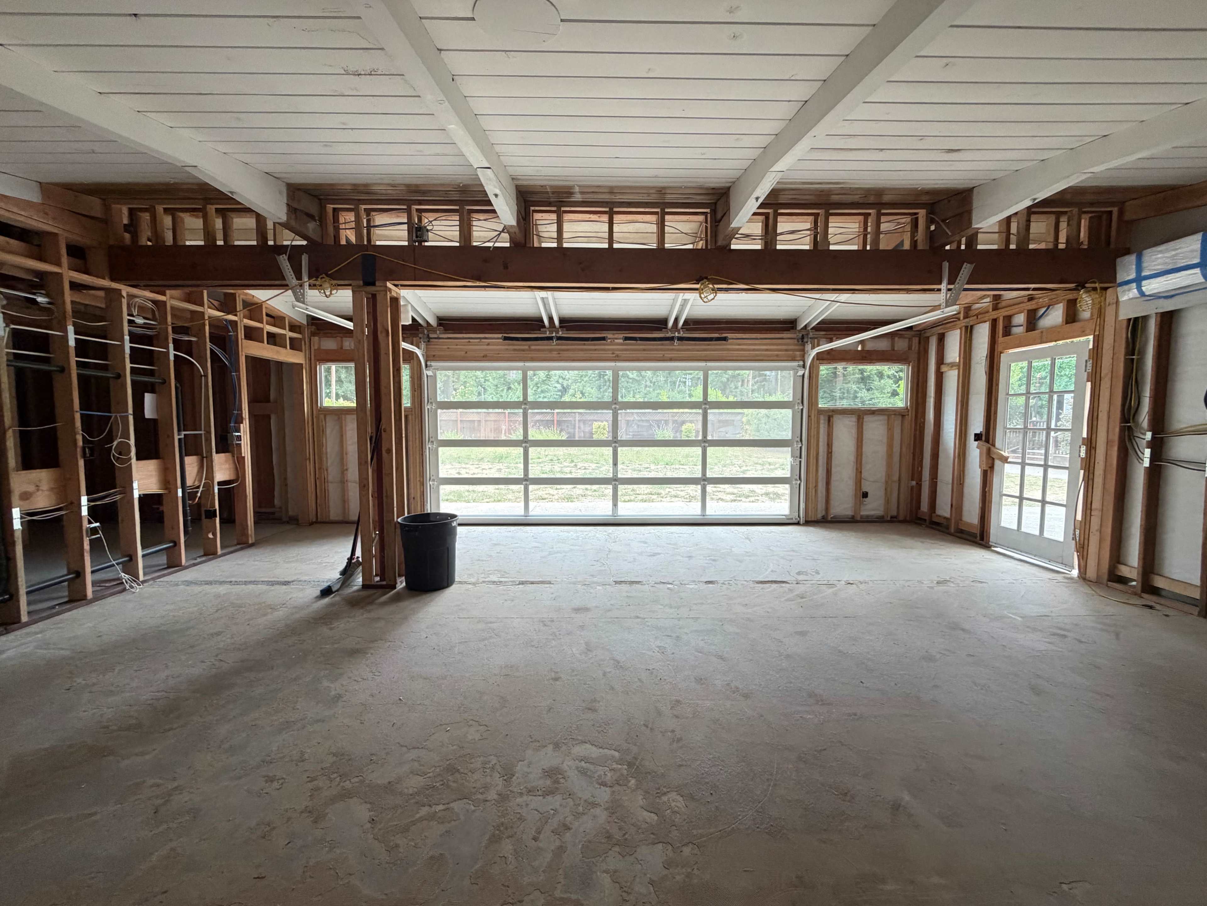 The image shows an unfinished interior space with exposed wooden beams, a large garage door in the back, and concrete flooring.