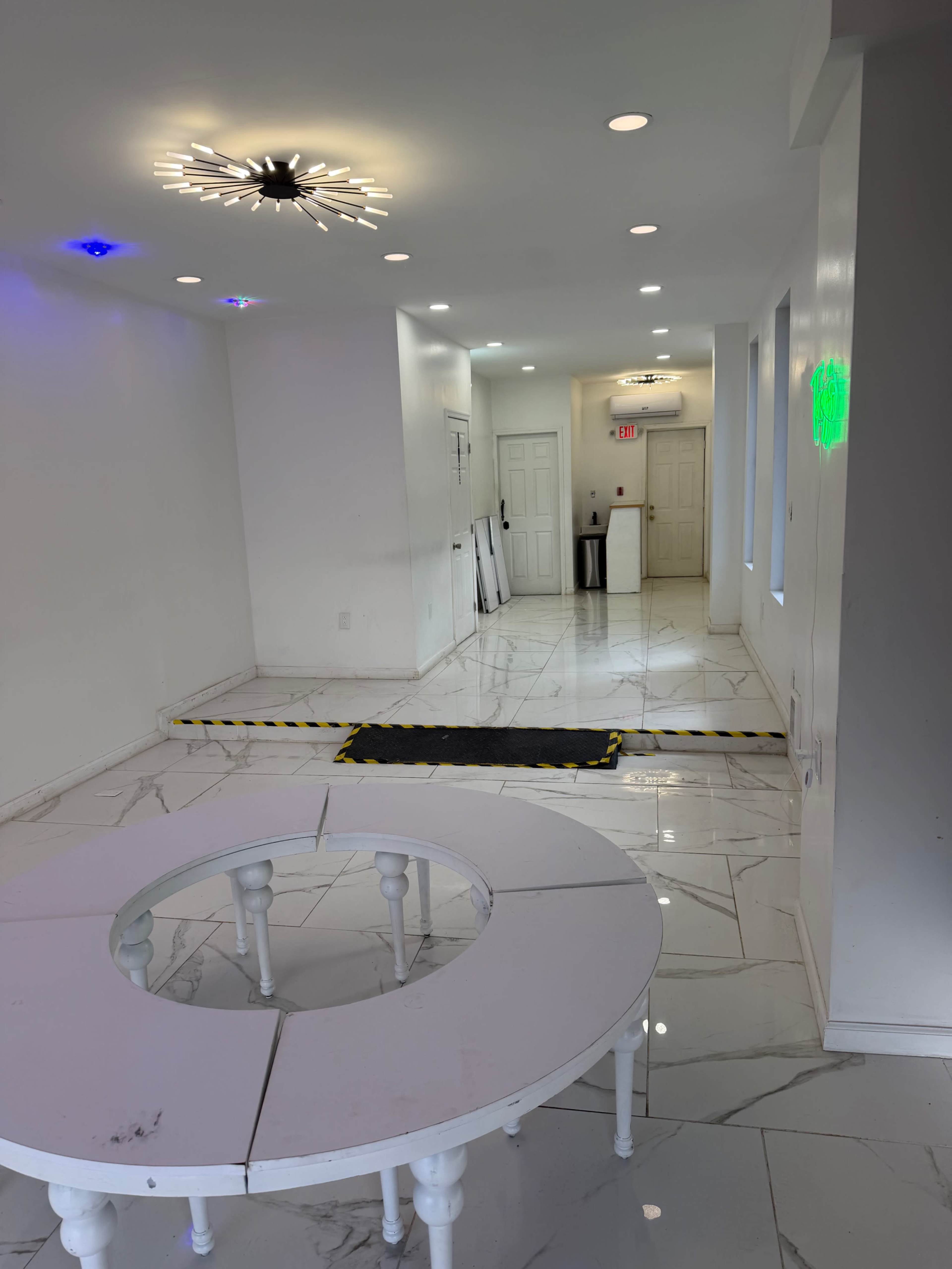 The image shows a spacious, brightly lit hallway with white walls, marble flooring, and circular tables arranged in the foreground.