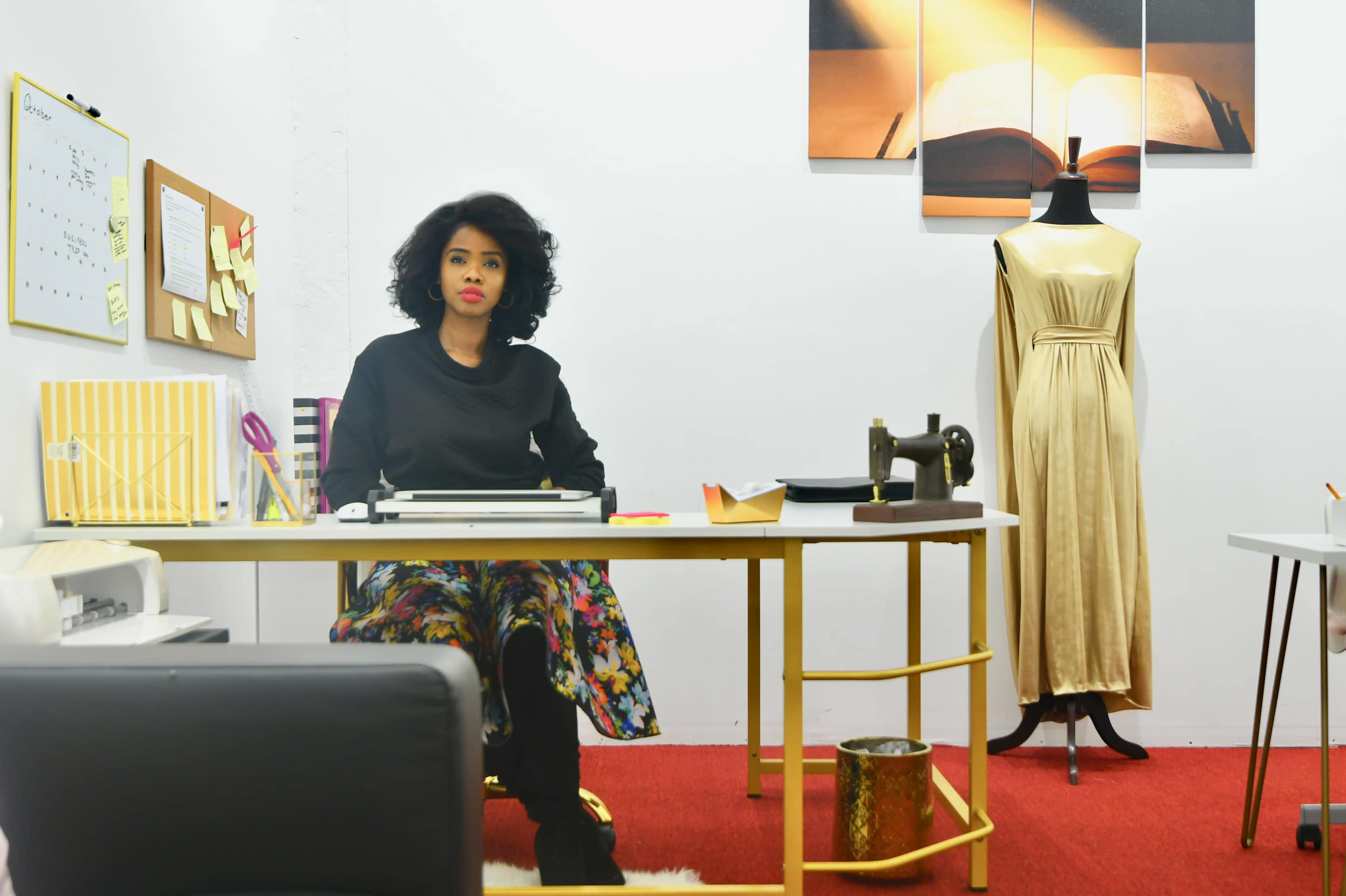 A woman with curly hair sits at a desk in a well-organized office featuring a sewing machine and a dress form beside her.