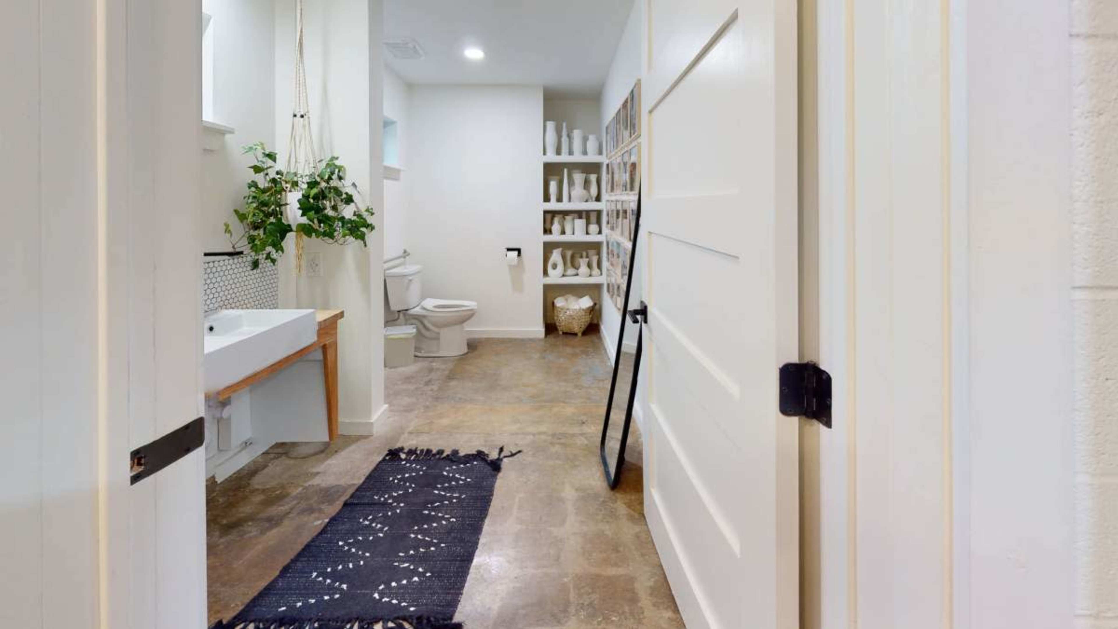 The image shows a hallway leading to a bathroom with a toilet, sink, and shelving unit, featuring a black rug on the floor and a hanging plant nearby.