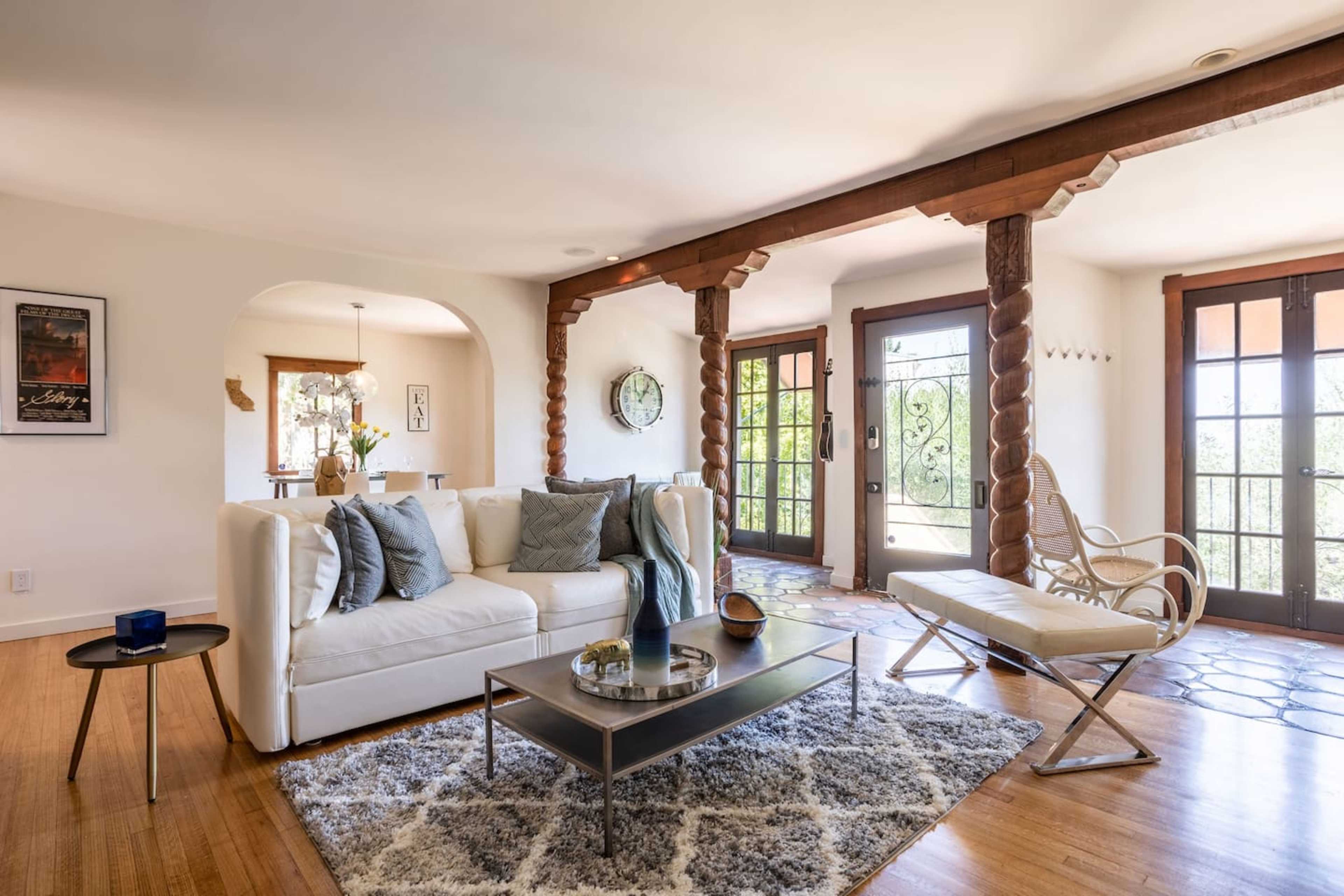 The image shows a living room with a white sofa, a glass coffee table, and large windows featuring wooden beams and a tiled floor.