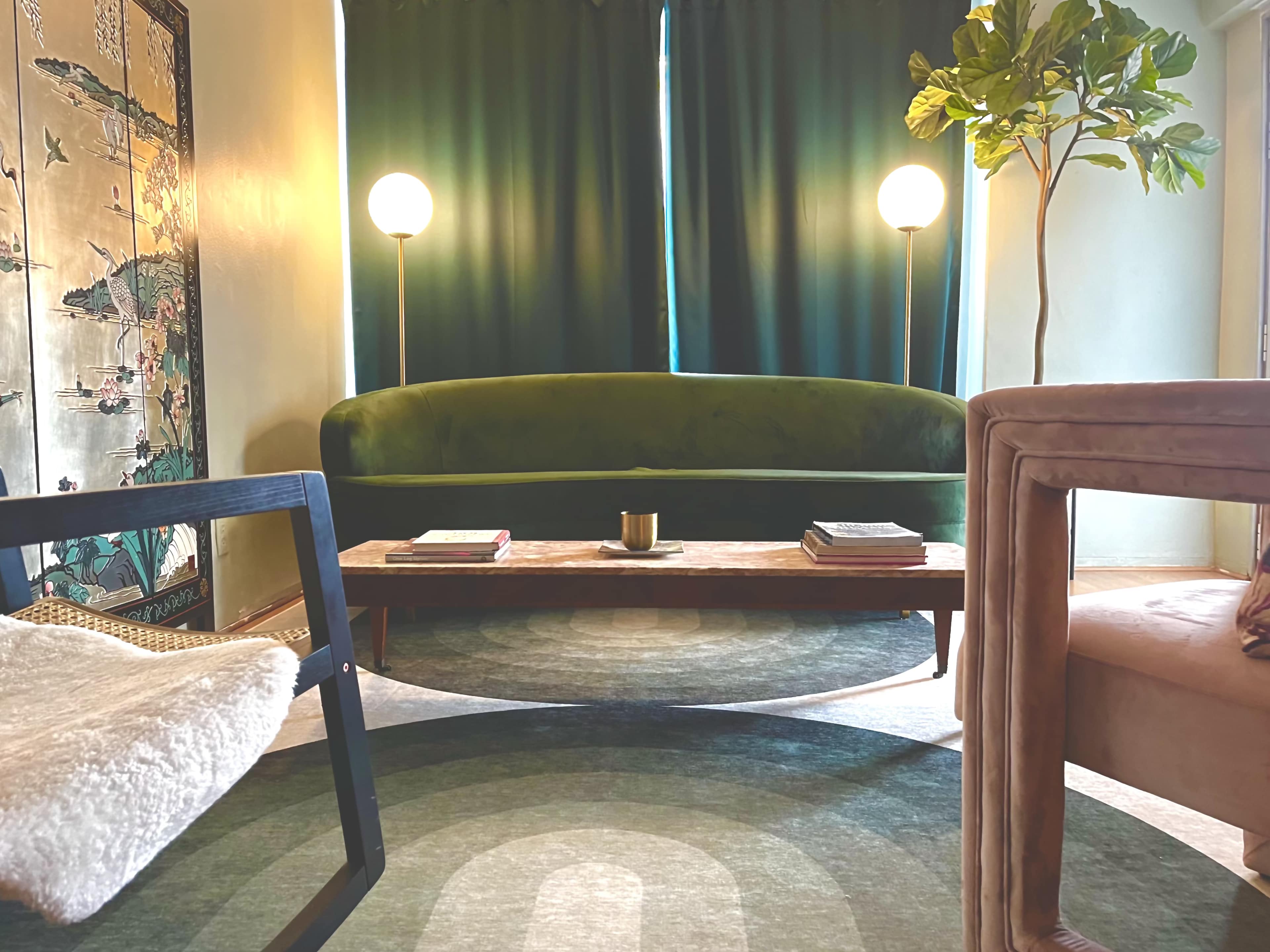 A cozy living room with a green curved sofa, a wooden coffee table, and two floor lamps, surrounded by dark curtains and a decorative wall panel.