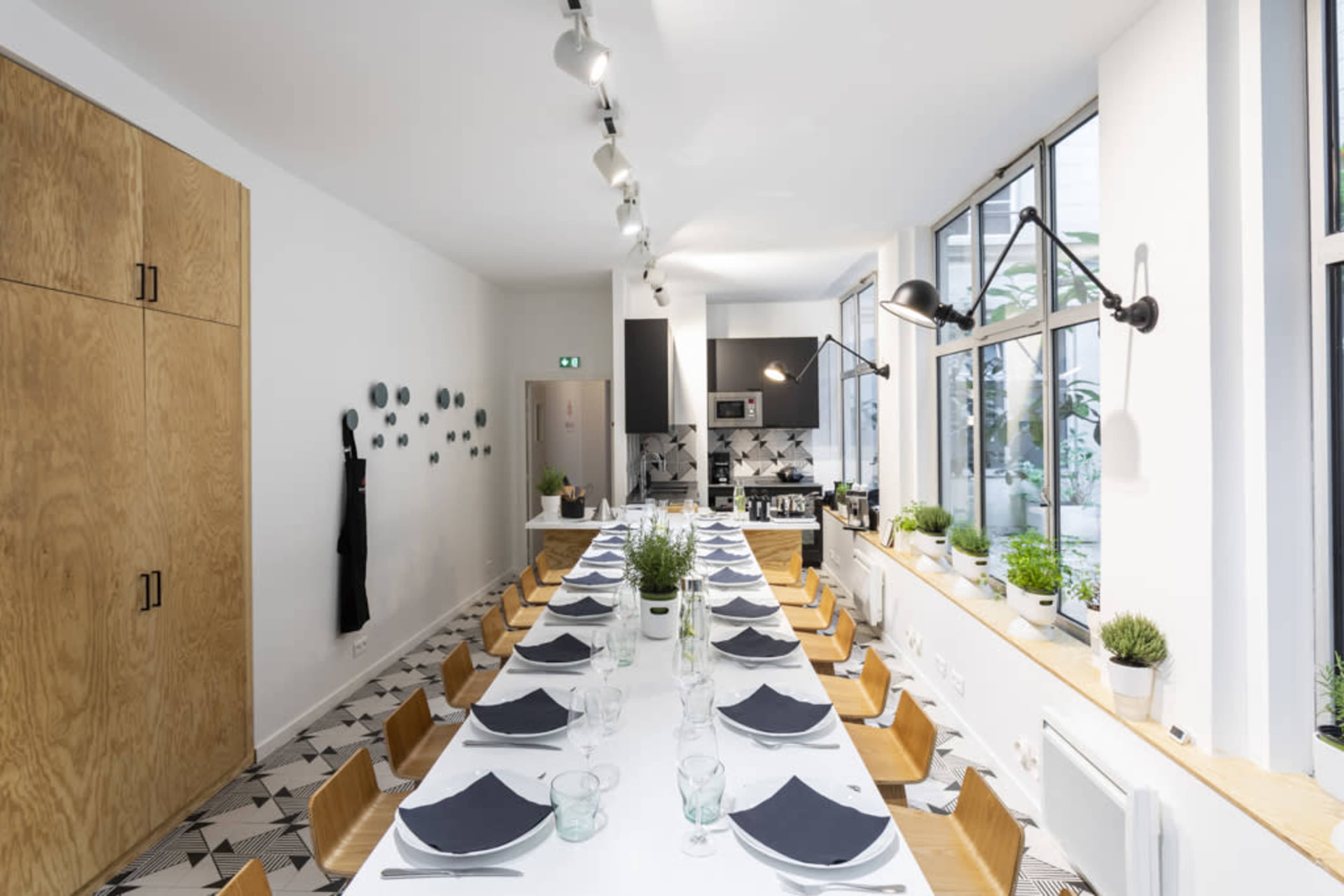 A long dining table is set with dark napkins and glassware in a bright, modern kitchen featuring large windows and minimalist decor.