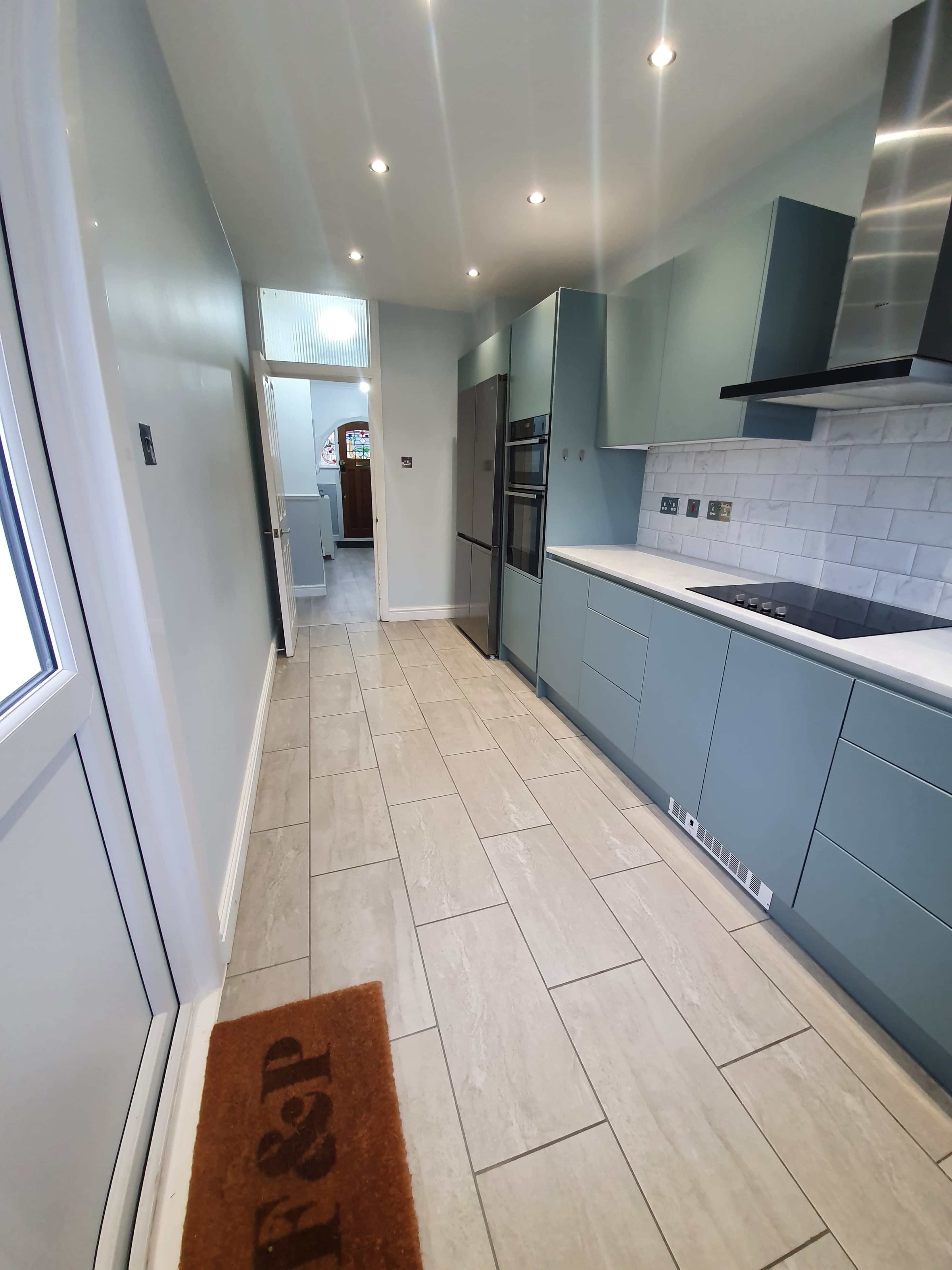 A modern kitchen with light blue cabinets, a white countertop, and a tiled floor, extending into a hallway at the far end.