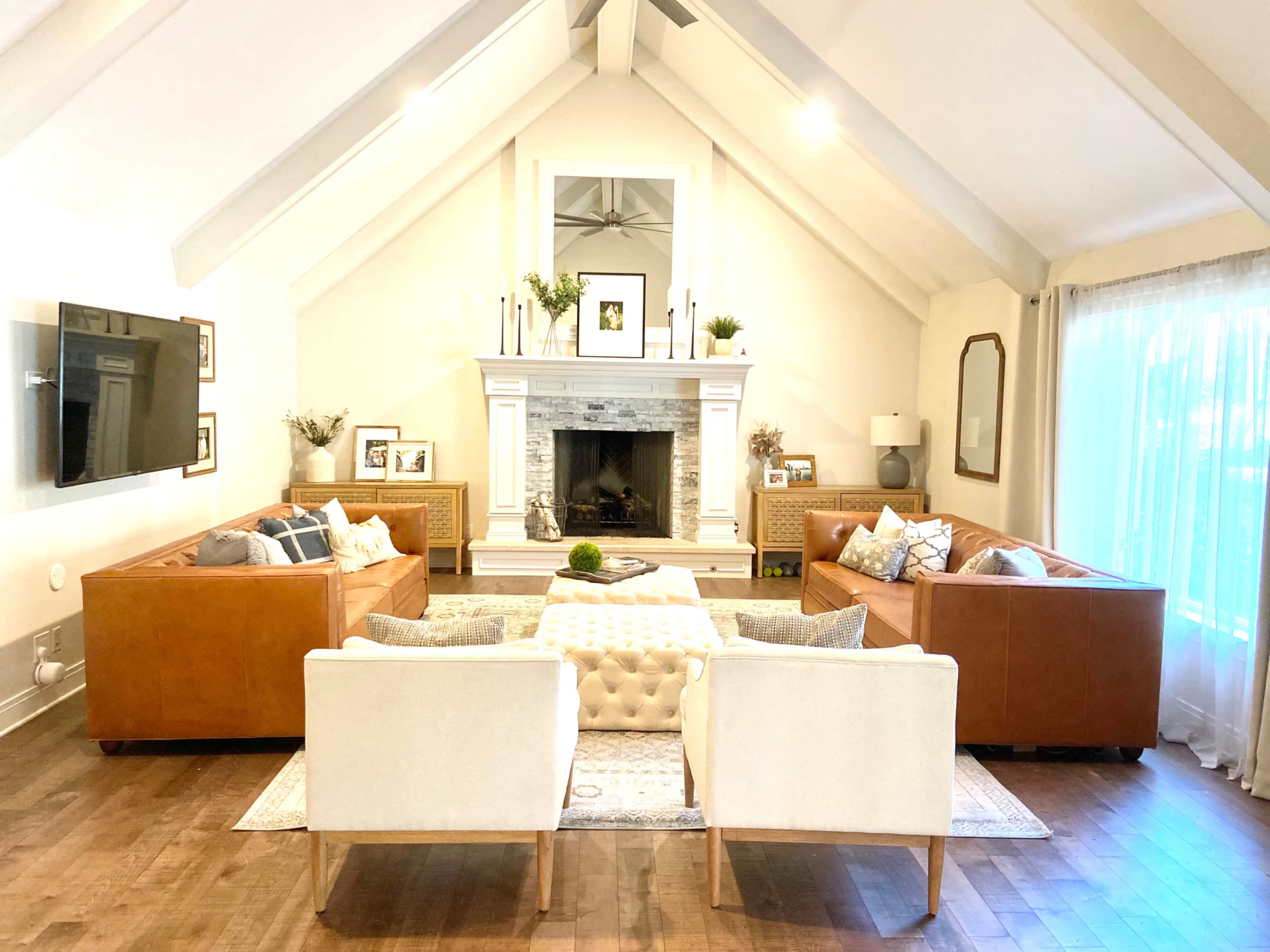 A spacious living room features tan leather sofas and white armchairs arranged around a central tufted coffee table in front of a stone fireplace.