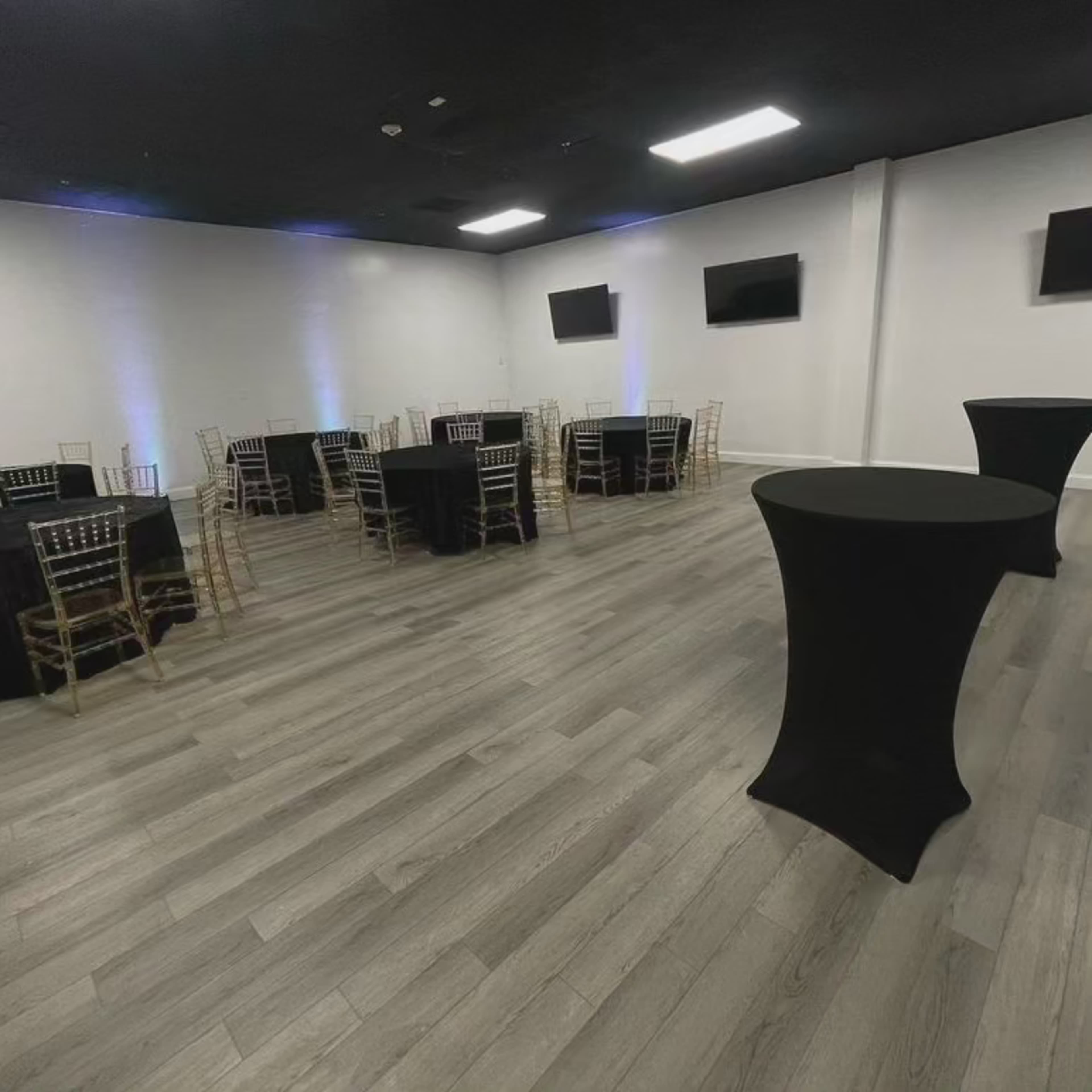 A spacious indoor event venue with several round tables covered in black tablecloths and two tall black cocktail tables.