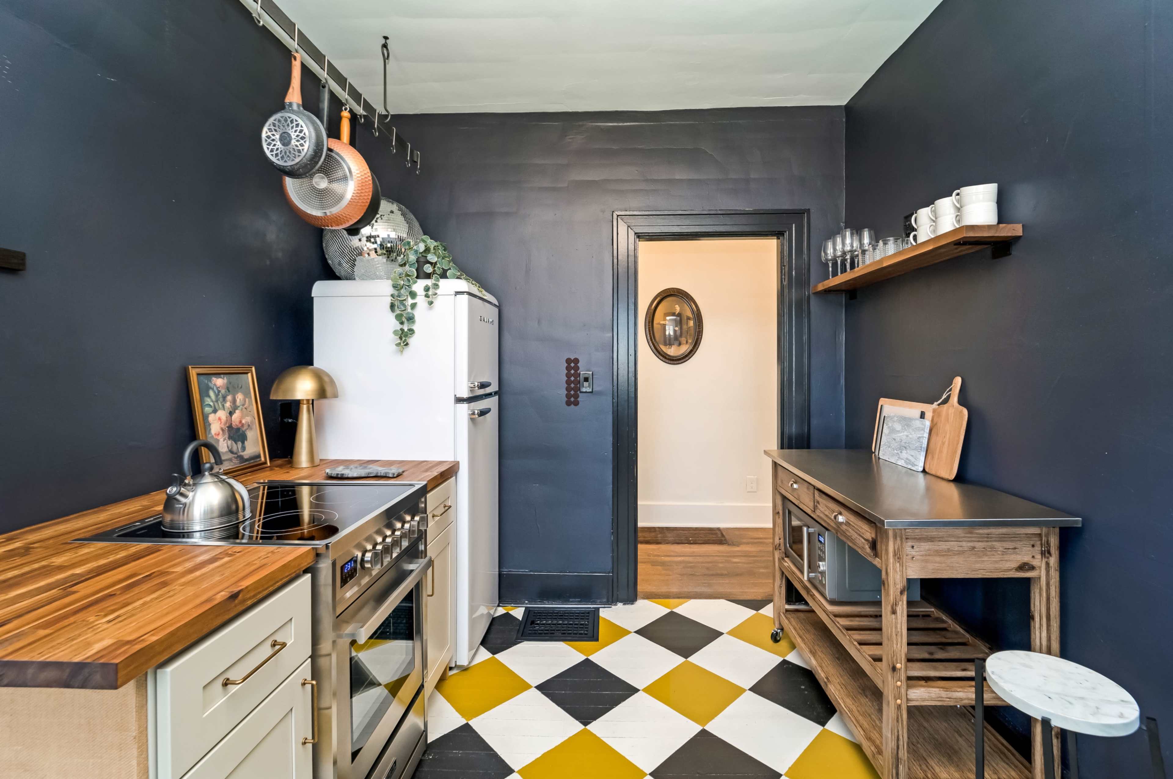 The image shows a kitchen with dark blue walls, a white refrigerator, a stainless steel stove, and a checkered yellow and white floor, leading to an adjacent room.