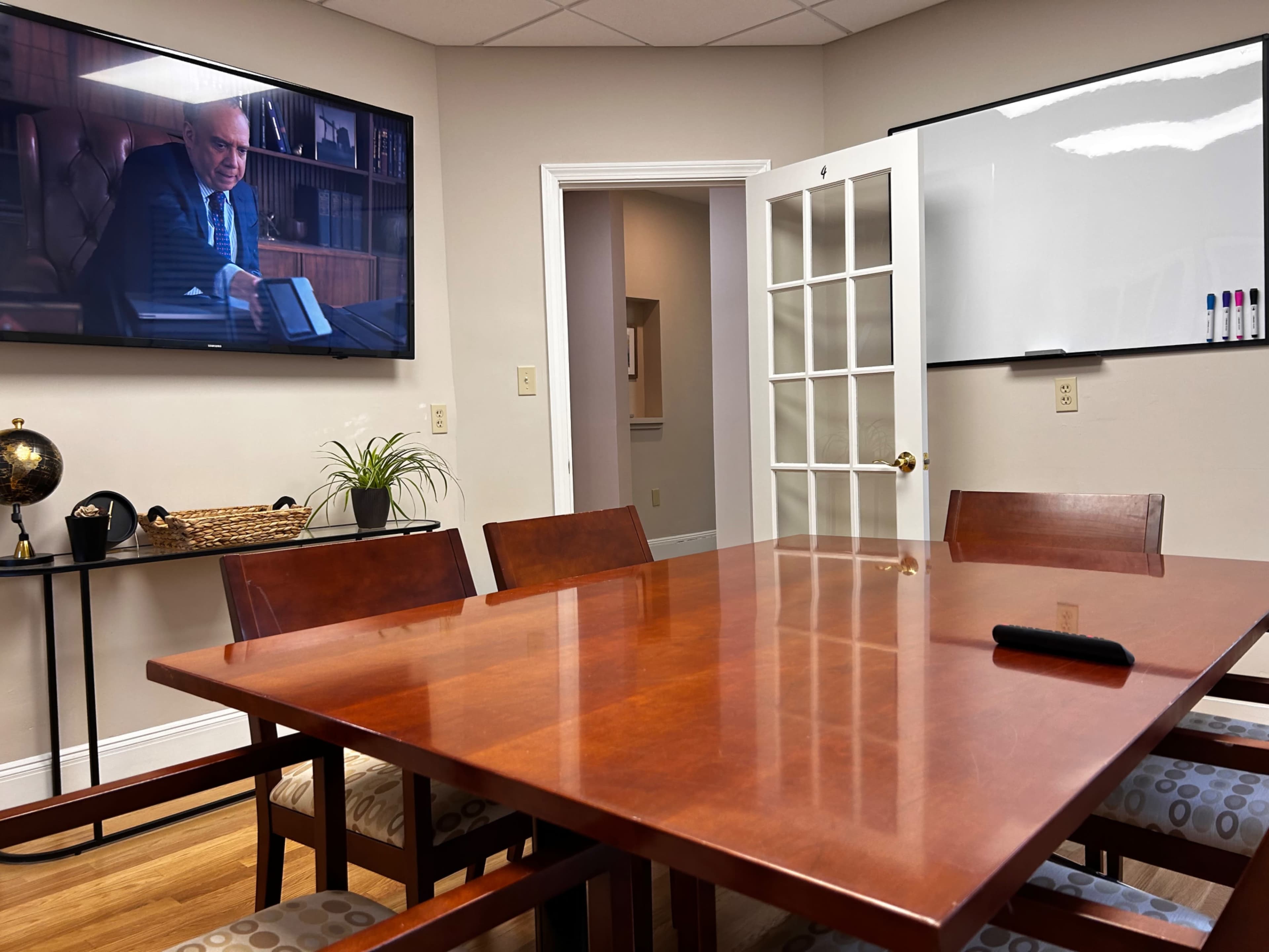 A conference room with a wooden table, chairs, a screen displaying a man, and a whiteboard on the wall.