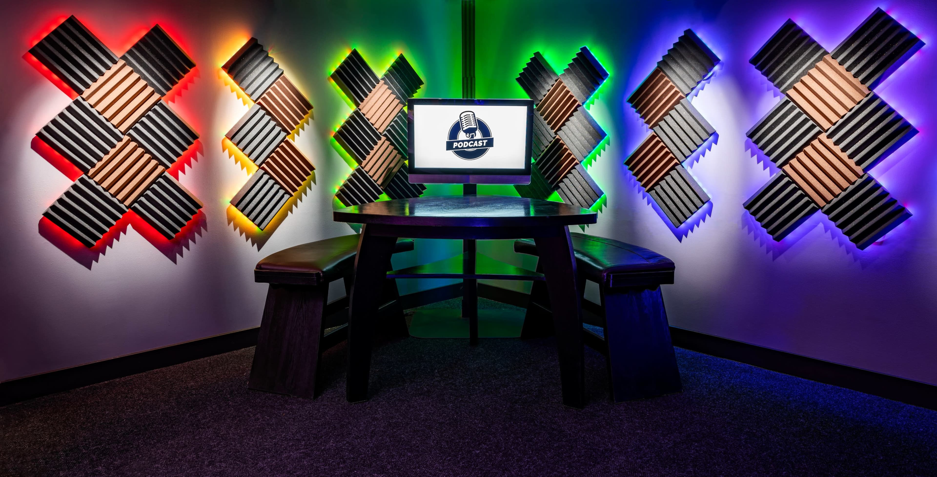 A podcasting studio features a central table with two benches, illuminated by colorful LED lights and surrounded by soundproofing panels.