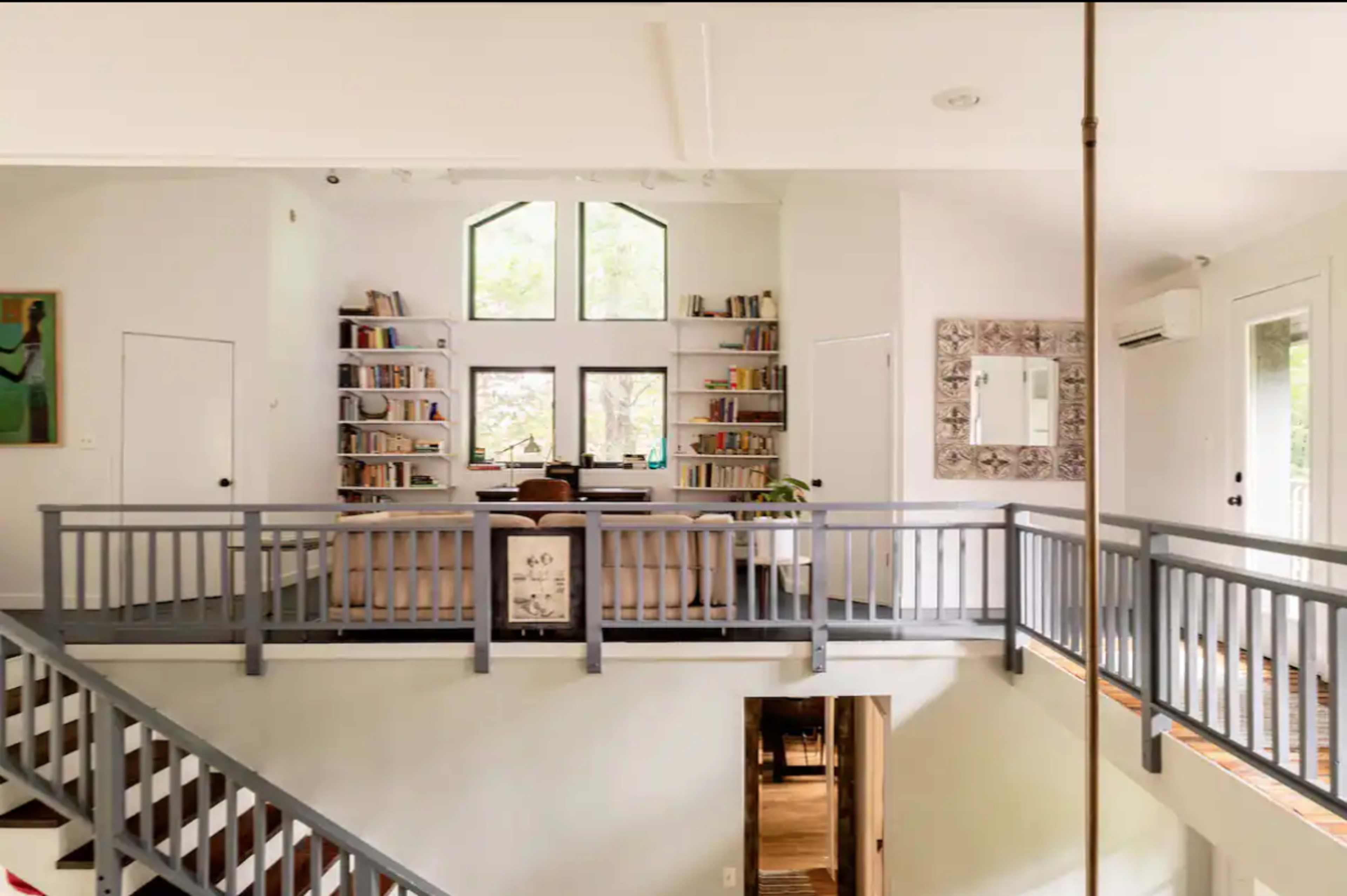 The image shows a two-level living space with a bookshelf-filled wall, a couch, and large windows allowing natural light to enter.