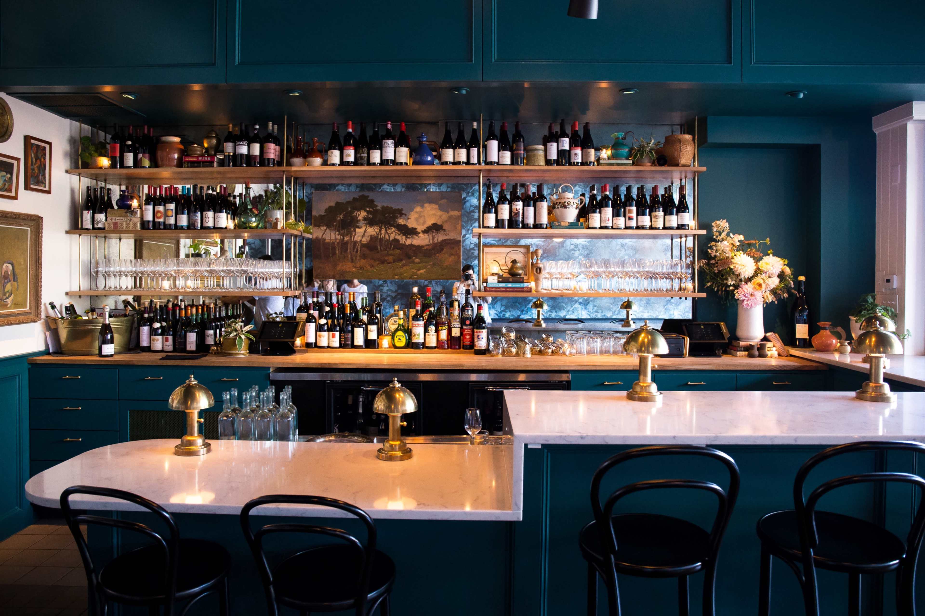 A stylish bar with teal cabinetry, shelves filled with an array of wine bottles, and two marble-topped counters with brass fixtures.