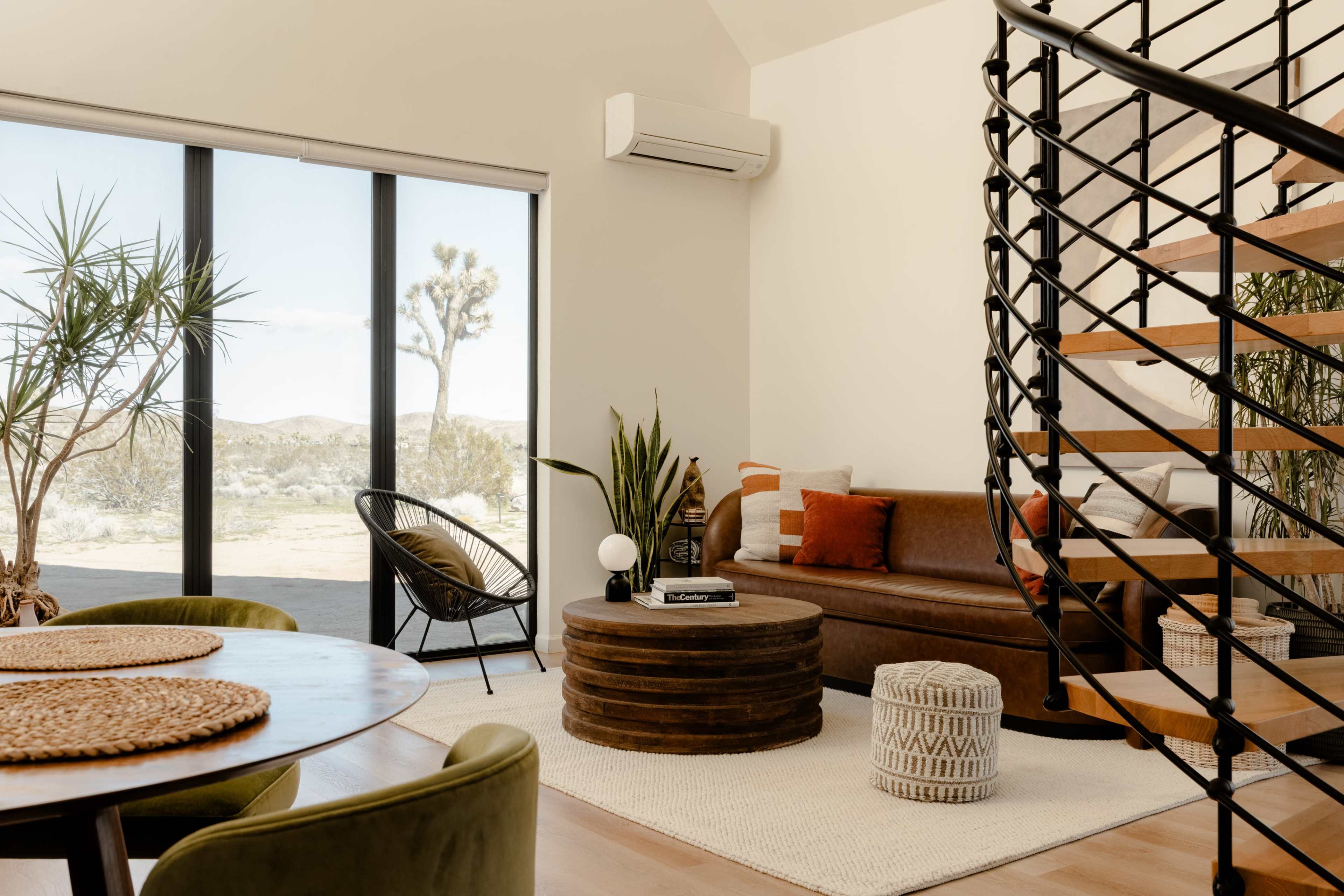 A modern living space features a leather sofa, round wooden coffee table, and a staircase, with large windows offering a view of a desert landscape.
