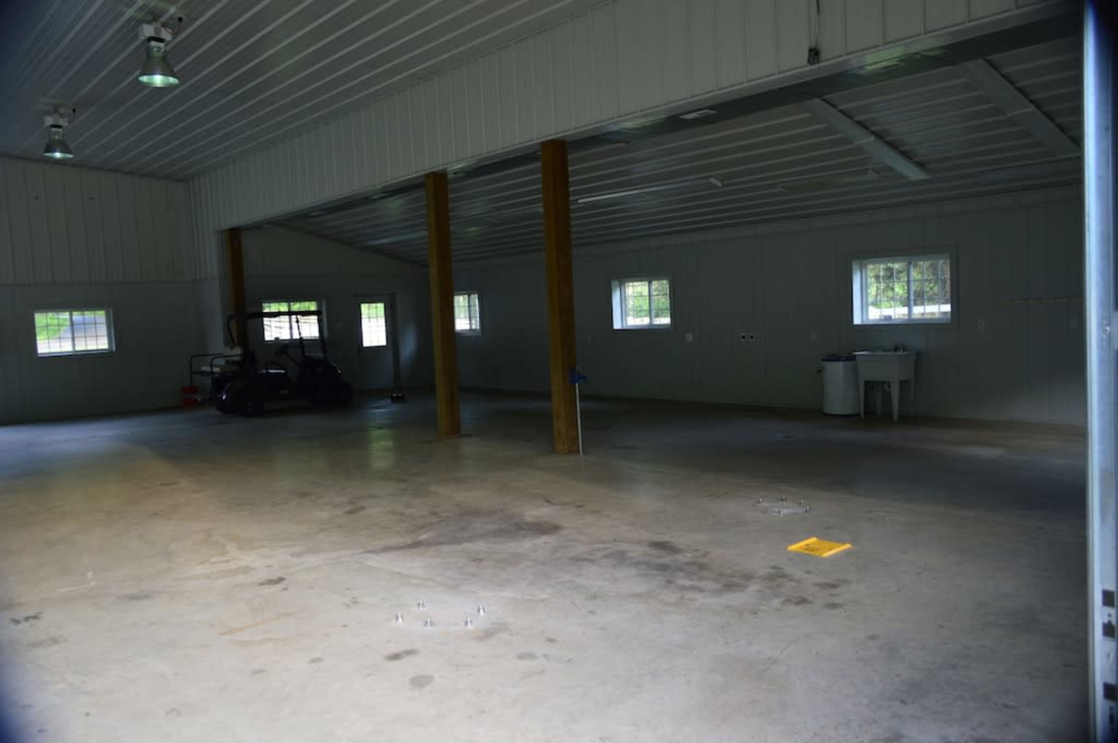 An empty indoor space with a concrete floor, metal walls, and wooden support beams, featuring a few windows and a golf cart in the background.