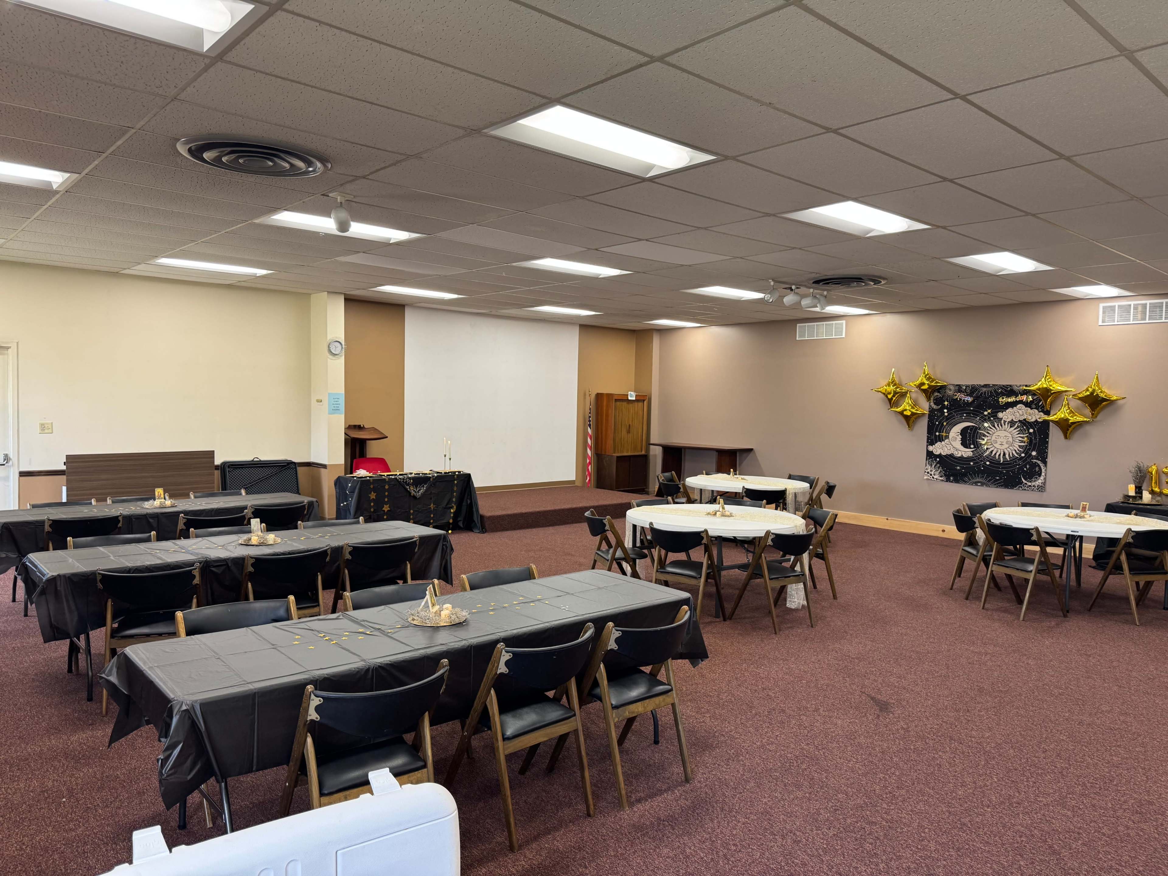 A spacious room is set up for an event with multiple tables covered in black cloths, gold decorations, and a projector screen at the front.