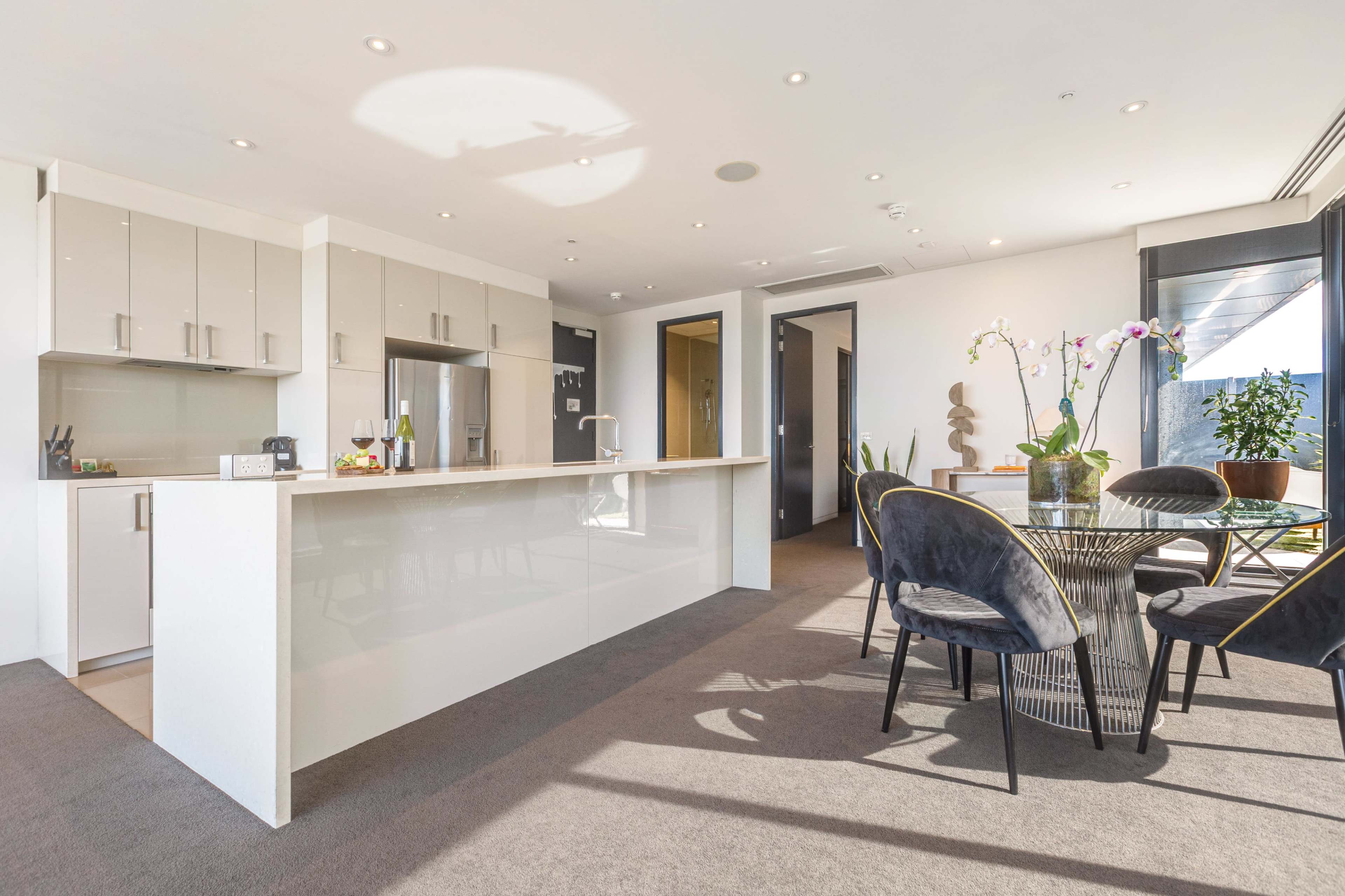 The image shows a modern kitchen and dining area with a sleek countertop, dining table, and large windows allowing natural light to fill the space.