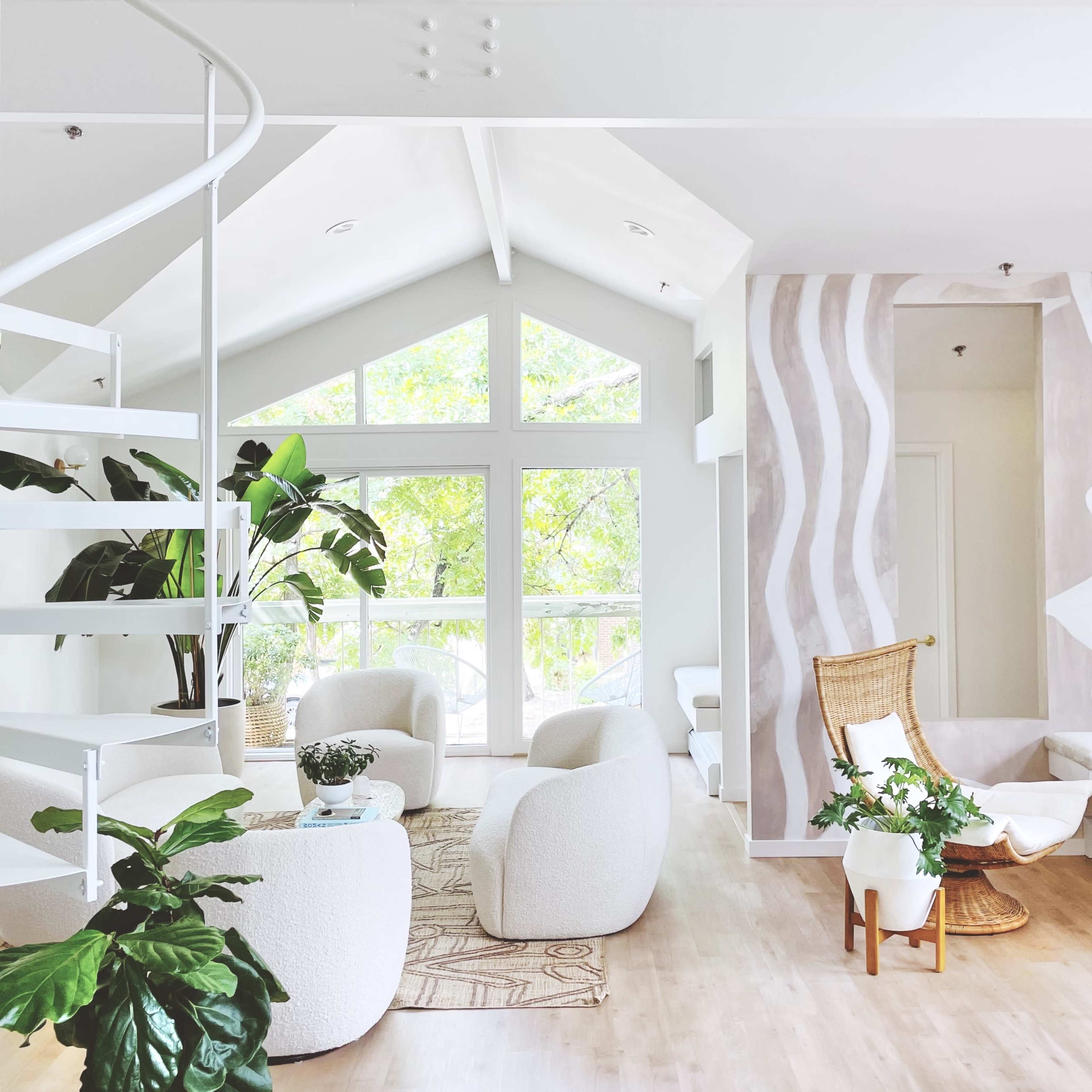 A bright, modern living space features a spiral staircase, plush white seating, large windows with natural light, and decorative plants.