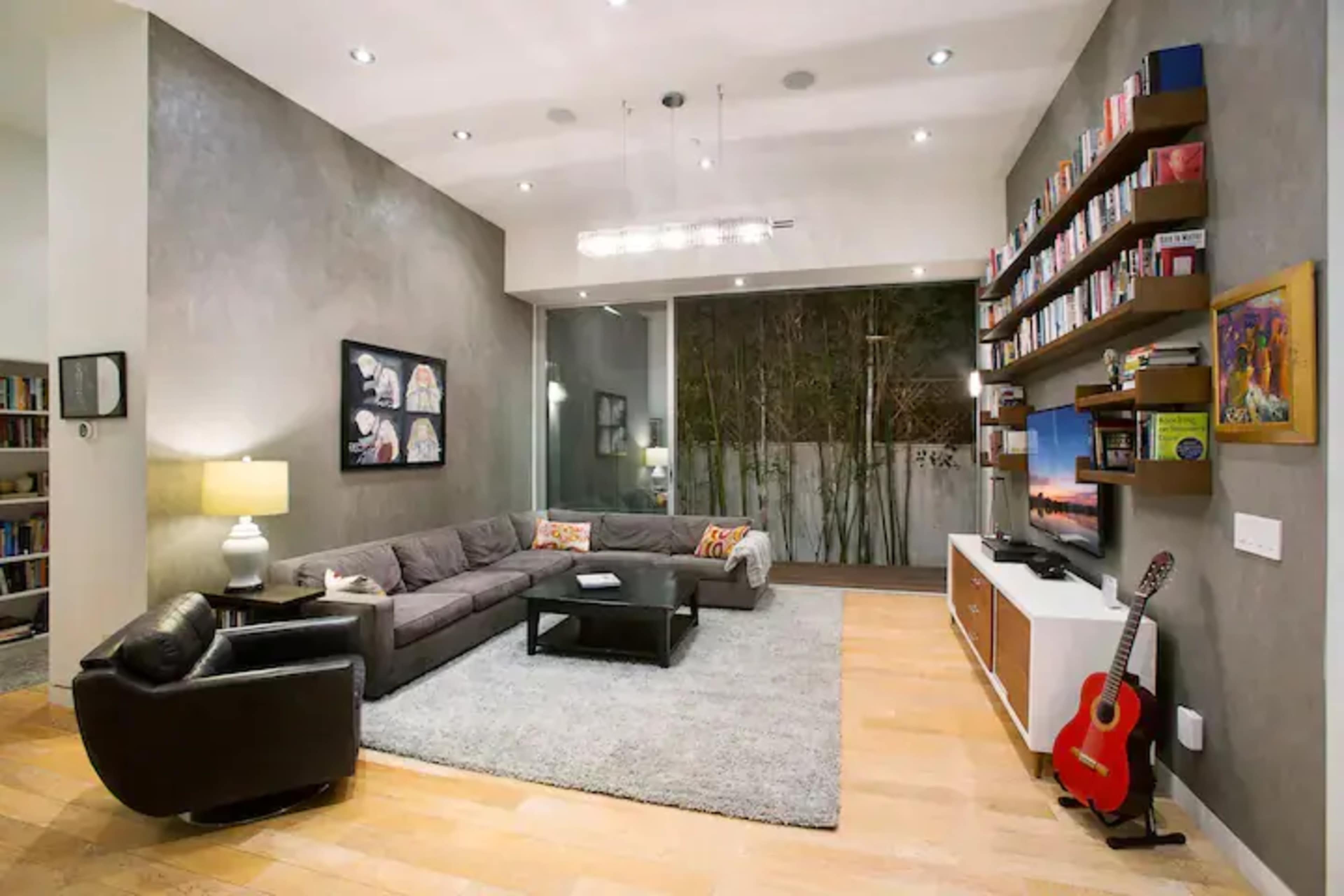 The living room features a gray sectional sofa, a black armchair, a coffee table, and a large bookshelf along one wall, with large windows revealing a bamboo garden outside.