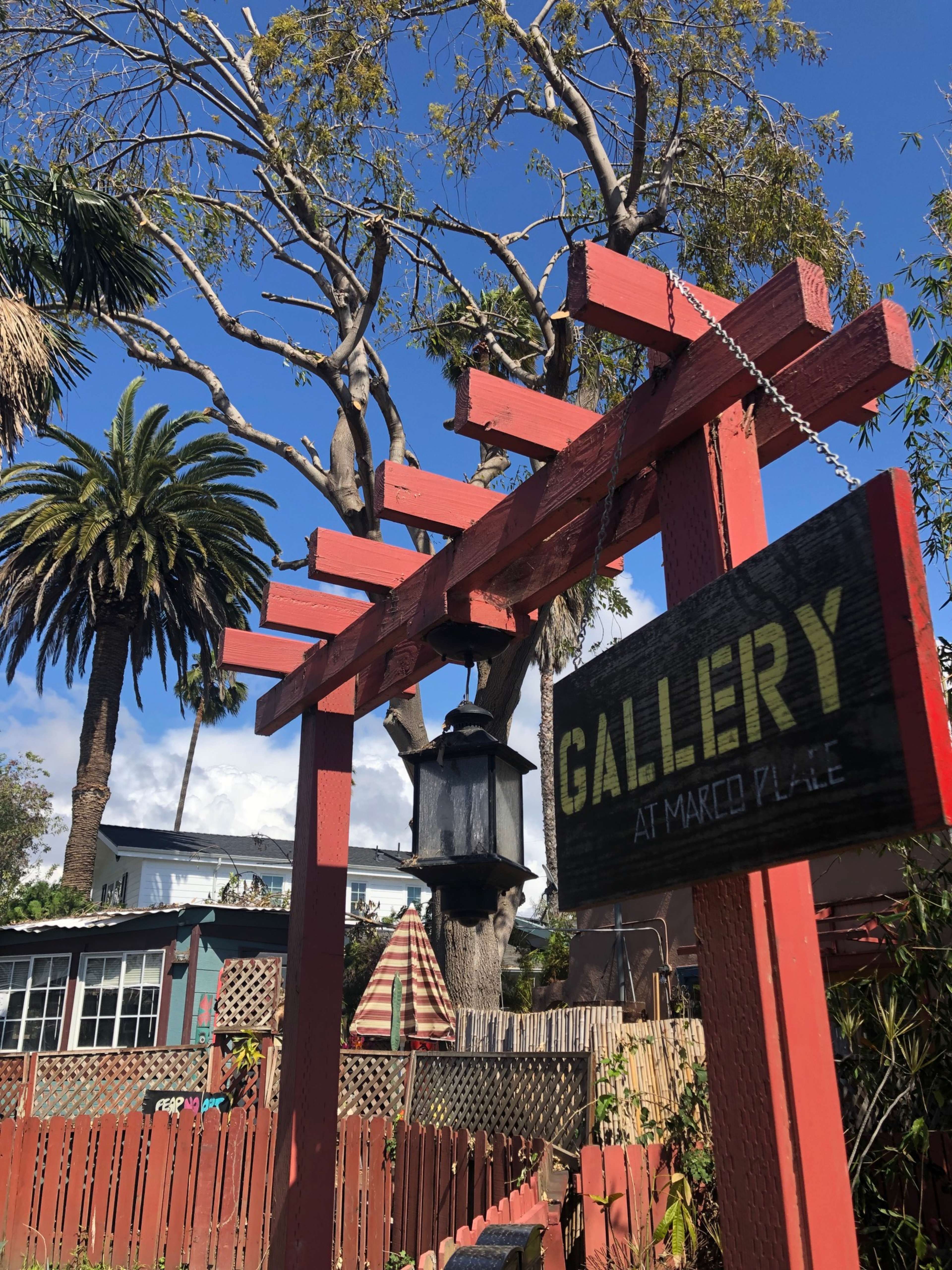 A wooden sign reading "GALLERY AT MARCO PLACE" hangs from a red trellis in a garden with palm trees and a cloudy sky in the background.