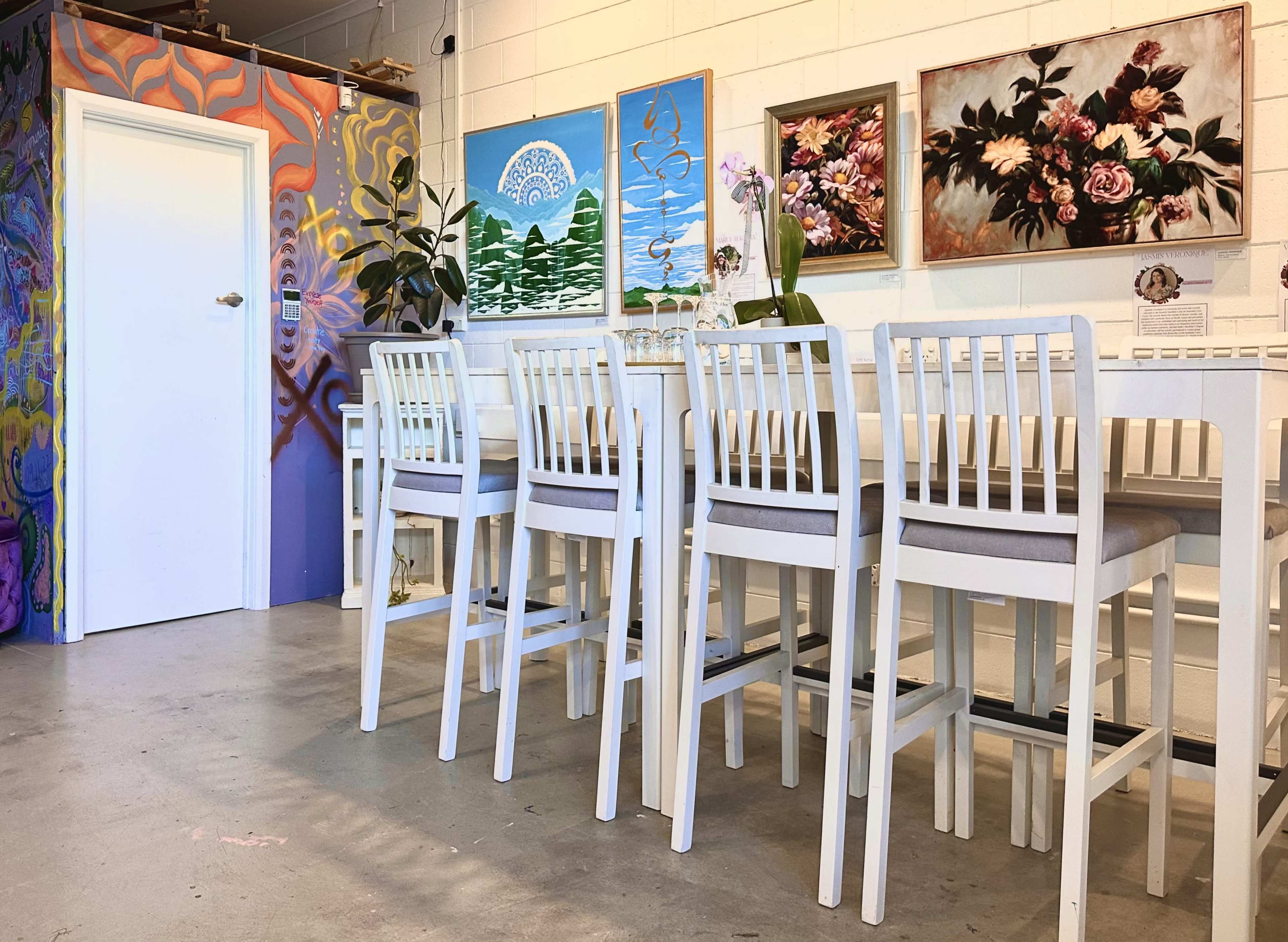 The image shows a brightly lit room with a long table and white high-backed chairs, decorated with various framed paintings on the walls.