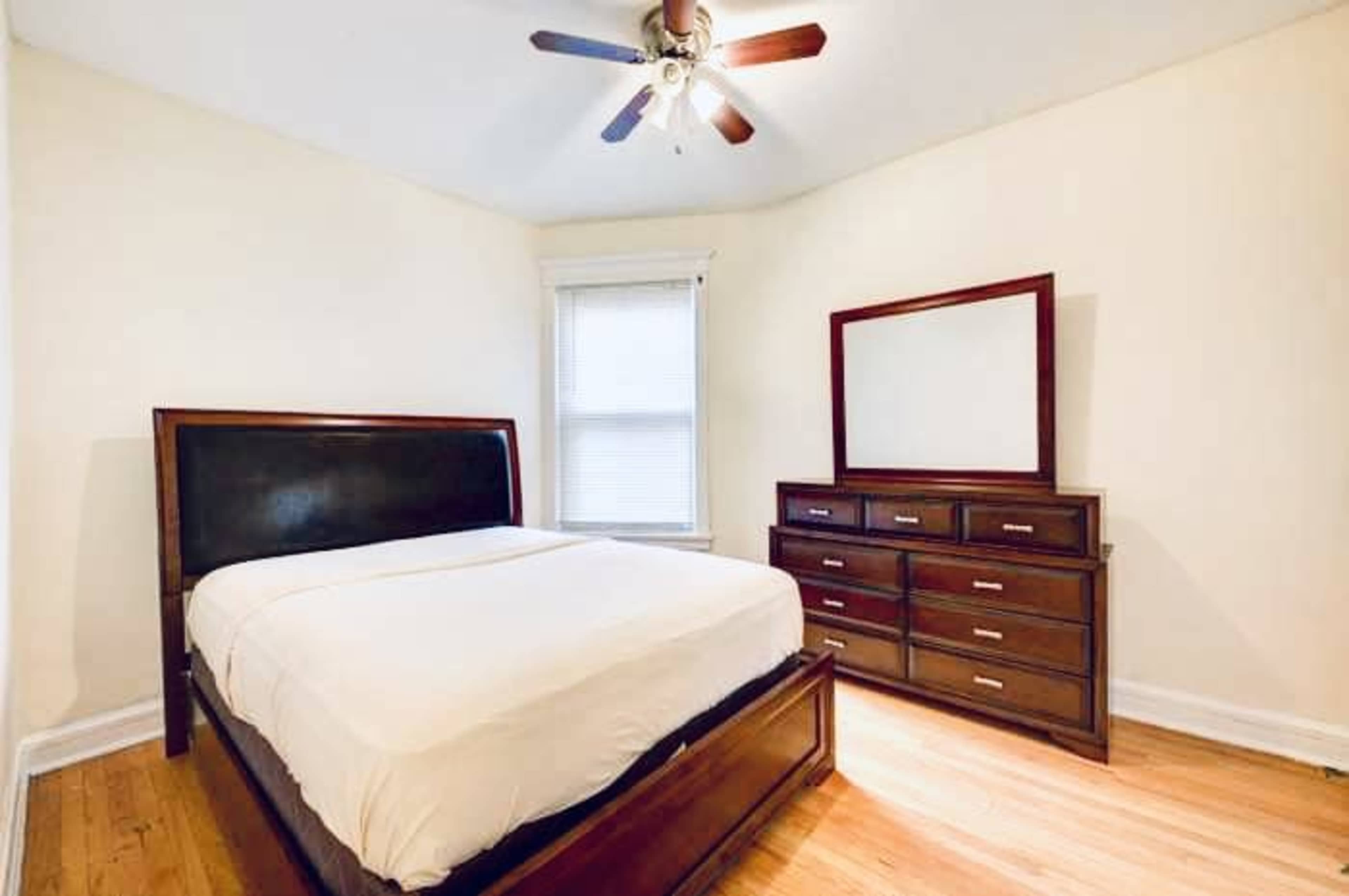 The room features a wooden bed frame with a white mattress, a brown dresser, and a mirror against the wall in a light-colored space.