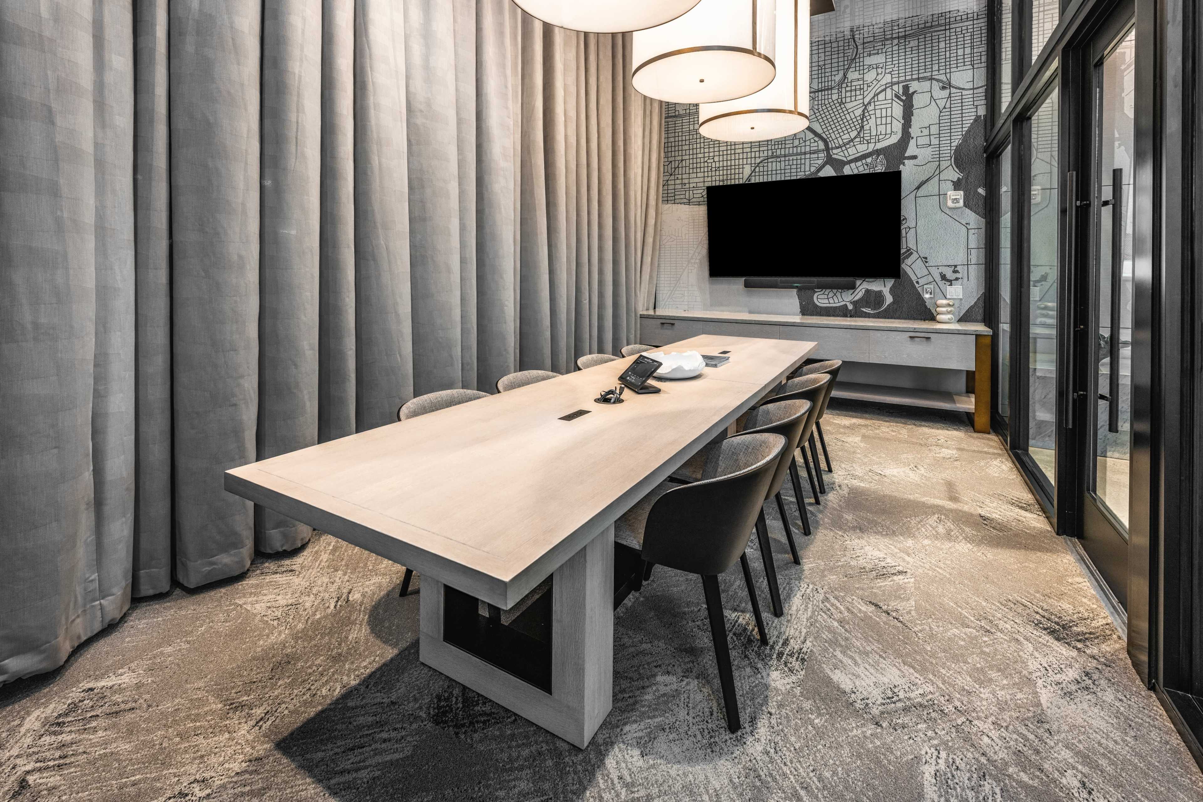 A long, wooden conference table is surrounded by eight chairs in a stylish meeting room with gray curtains and a large wall-mounted screen.