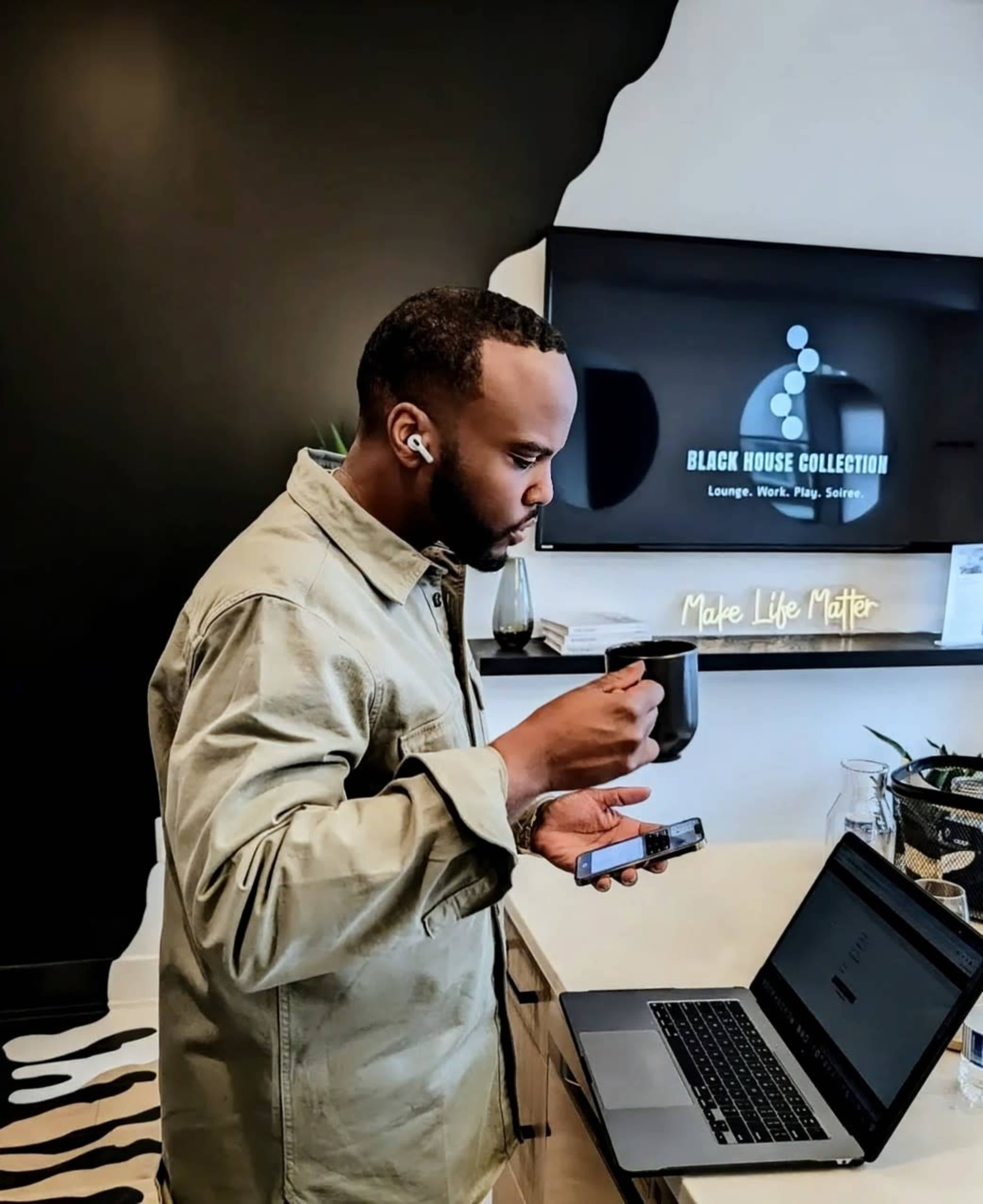A man is standing in an office environment, holding a mug in one hand while using a smartphone with the other, next to a laptop on a desk.