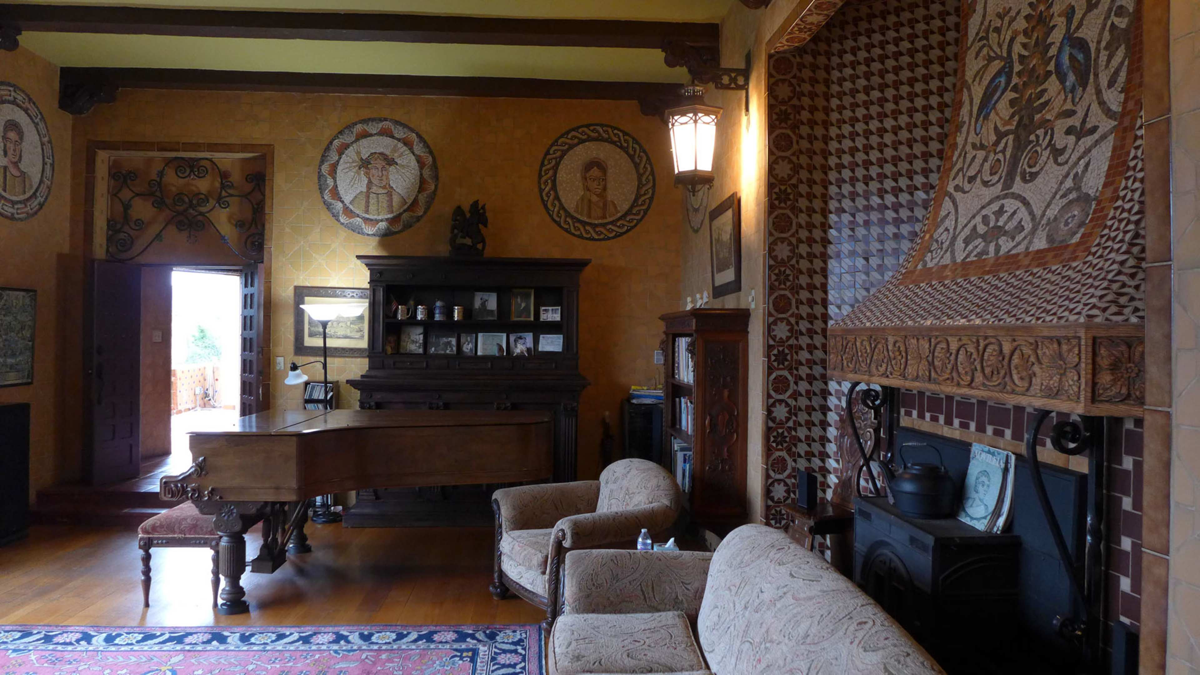 The image shows a room featuring a grand piano, two upholstered chairs, a decorative fireplace with intricate mosaics, and wall murals depicting circular designs.