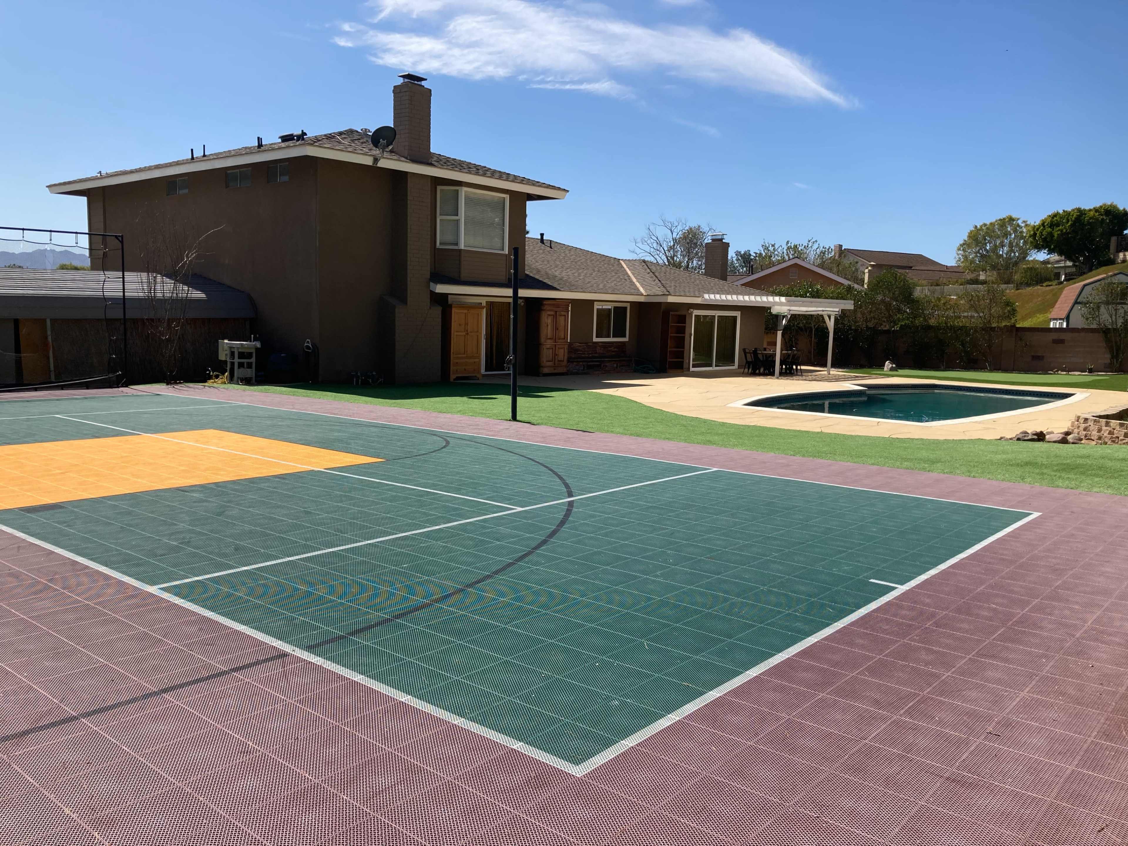 A basketball court in the foreground, with a swimming pool and a two-story house visible in the background.