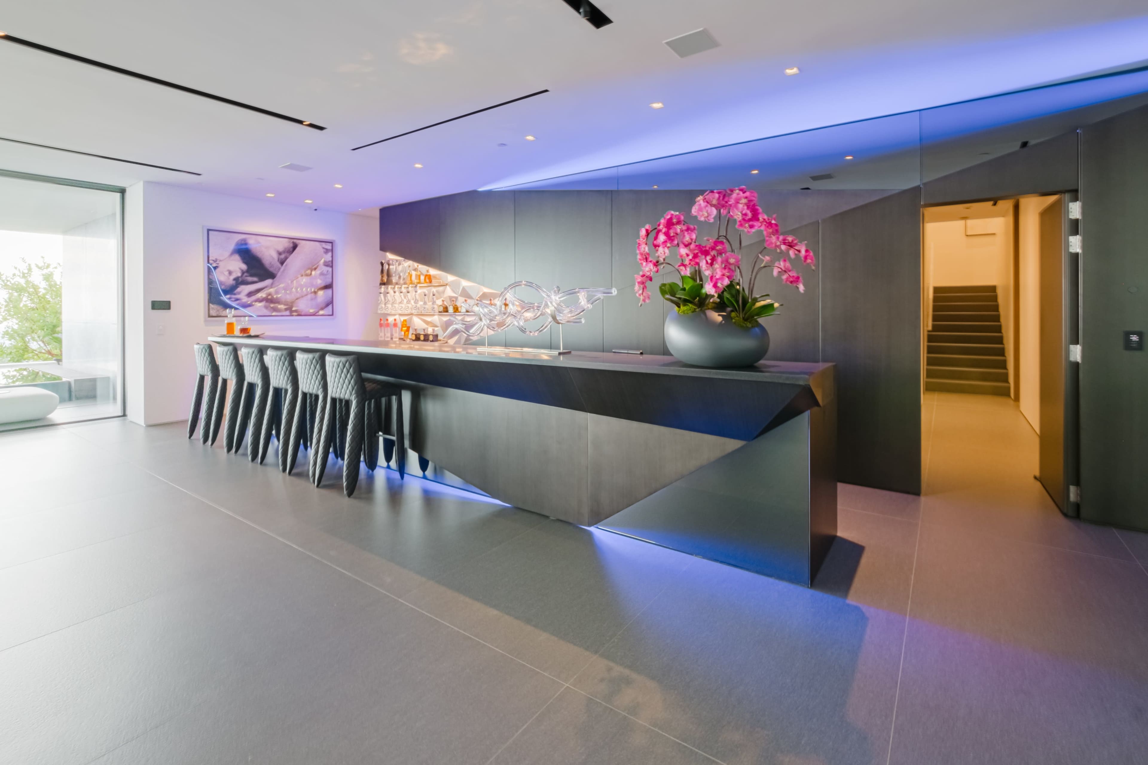 A modern kitchen features a sleek bar with high stools, illuminated by ambient lighting and adorned with a large vase of pink orchids.
