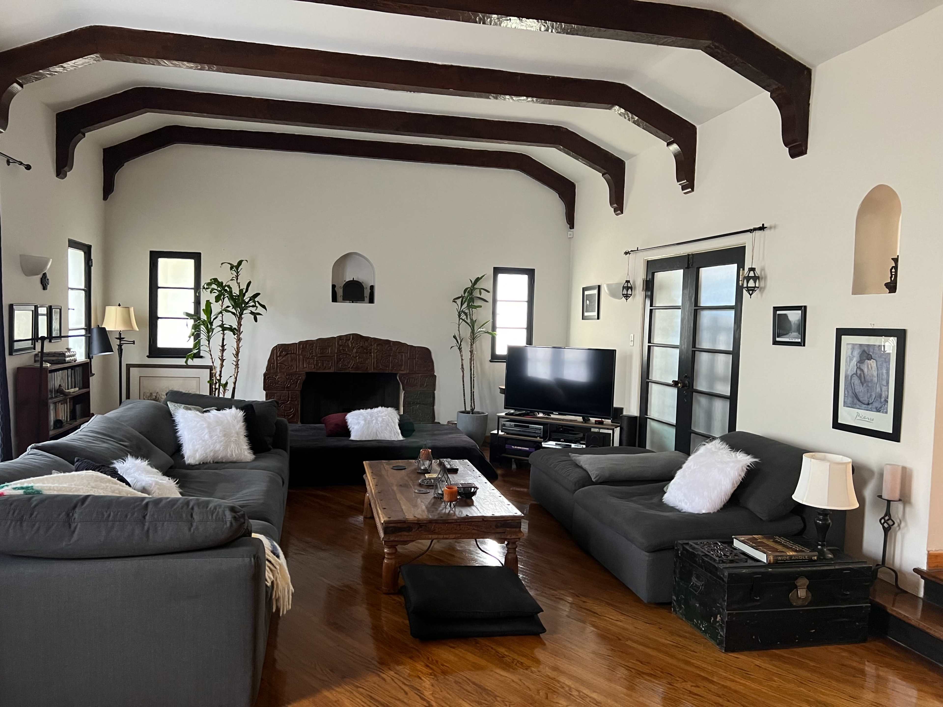 The image shows a spacious living room with vaulted ceilings, wooden beams, seating arrangements around a coffee table, and a television set against a wall.