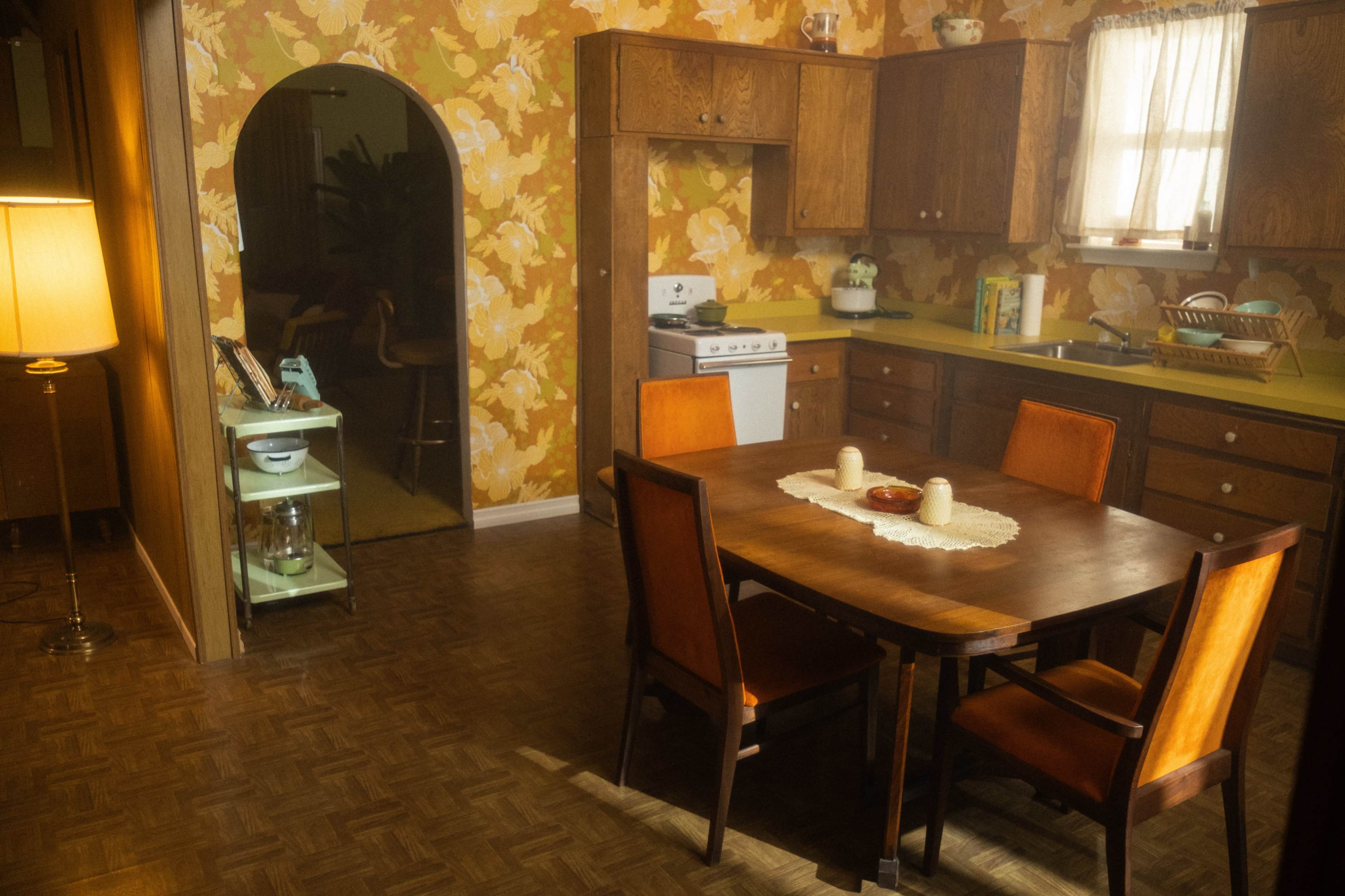 A wooden dining table is set with candles in a vintage kitchen featuring floral wallpaper and brown cabinetry.