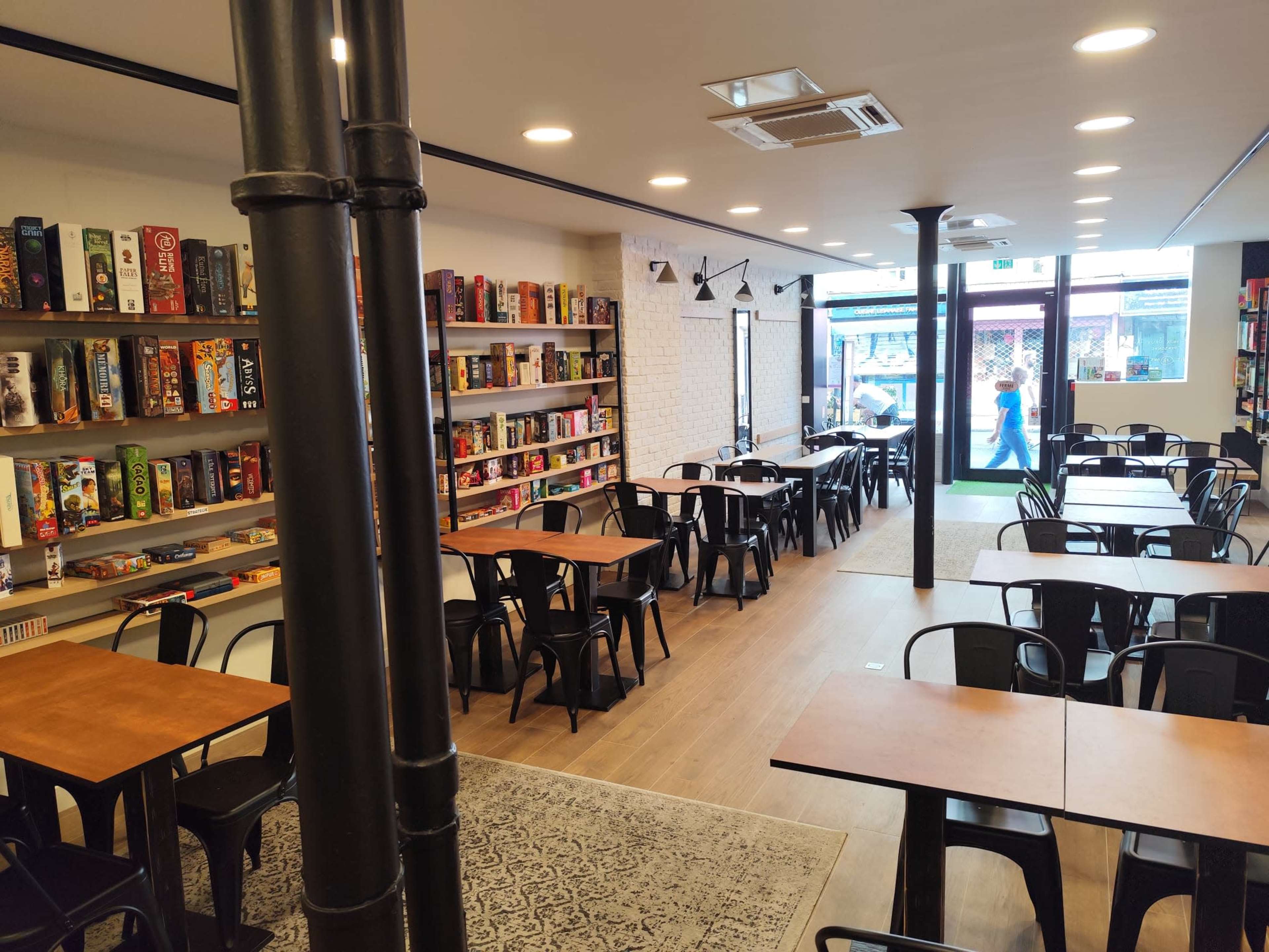 The image shows a spacious board game café with wooden tables and chairs, shelves lined with various board games, and a large window providing natural light.