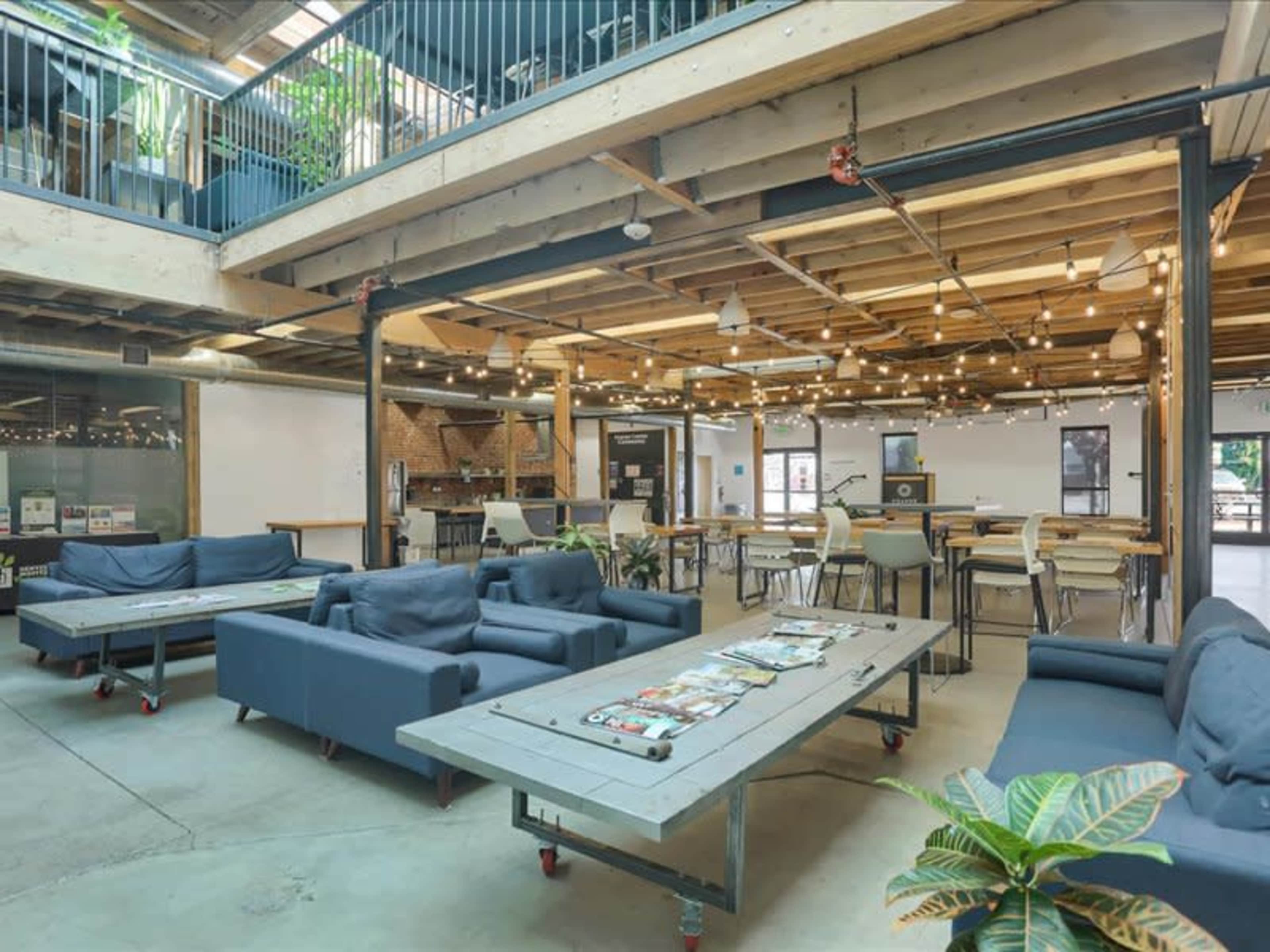 The image shows a modern open-concept workspace with multiple seating areas, wooden tables, and overhead lights, featuring a mix of couches and dining tables.
