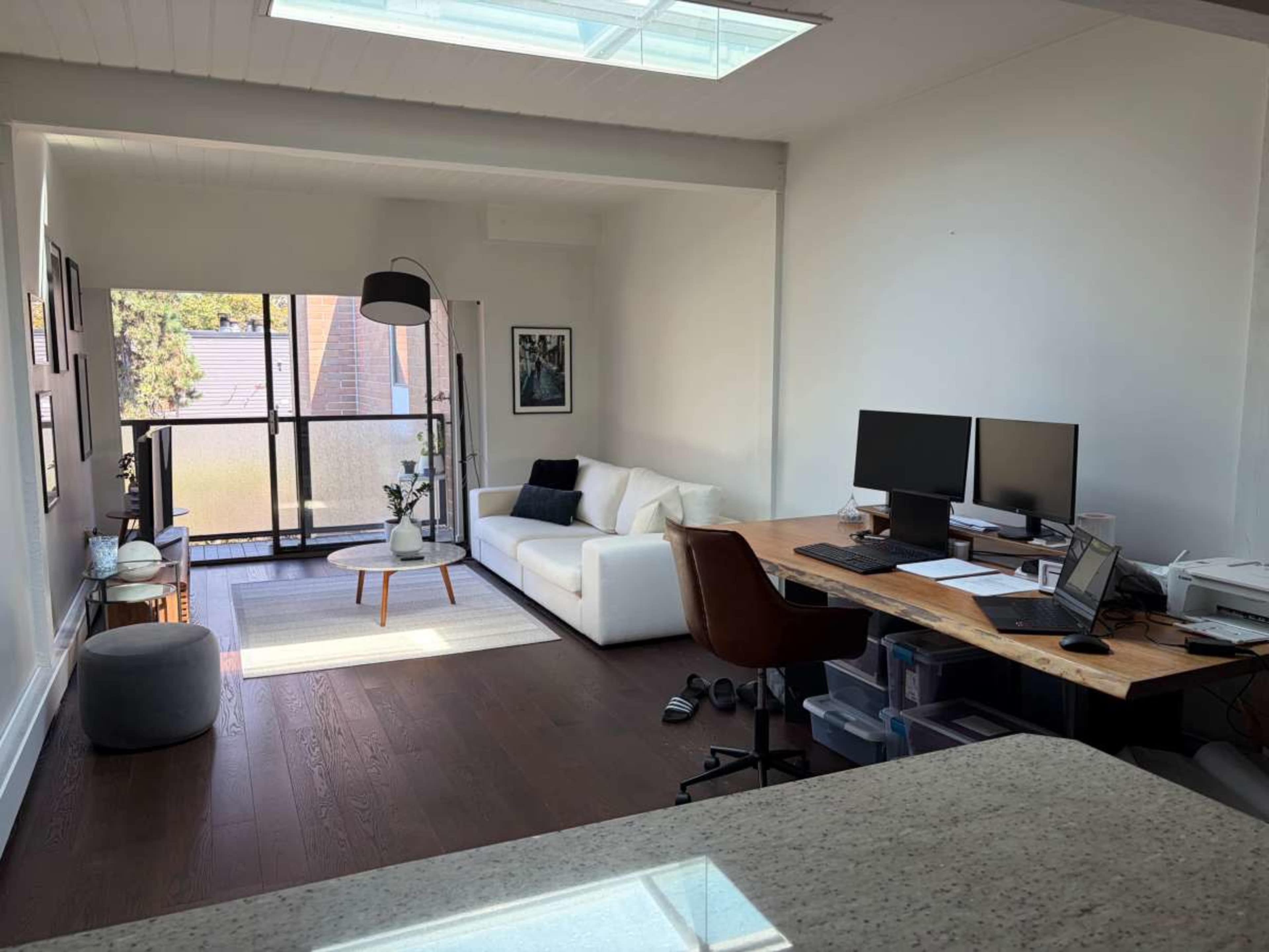 The image shows a modern living space featuring a white sofa, a coffee table, two computer monitors on a wooden desk, and large windows providing natural light.
