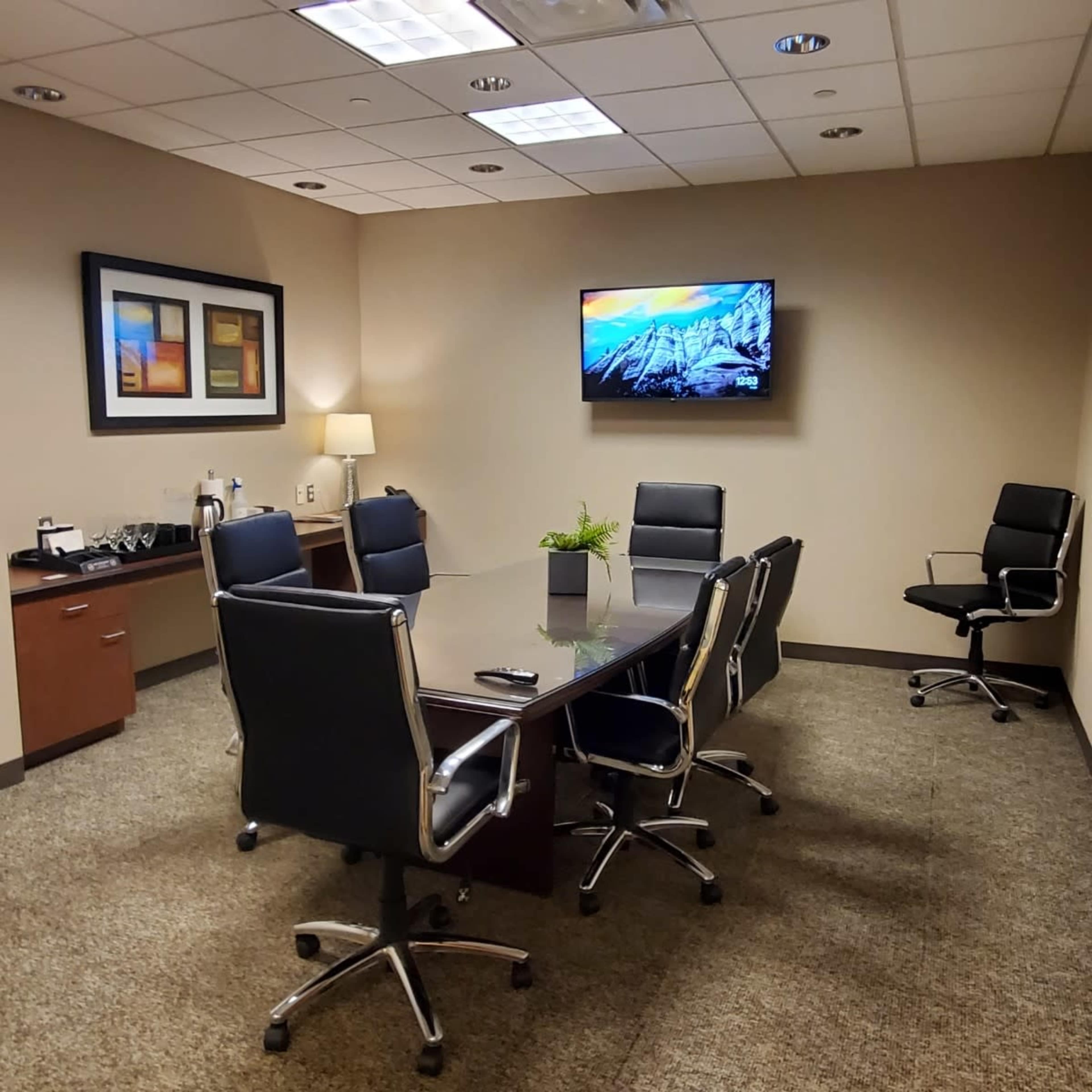 A conference room features a long rectangular table surrounded by black rolling chairs, a television mounted on the wall, and a small side table with refreshments.